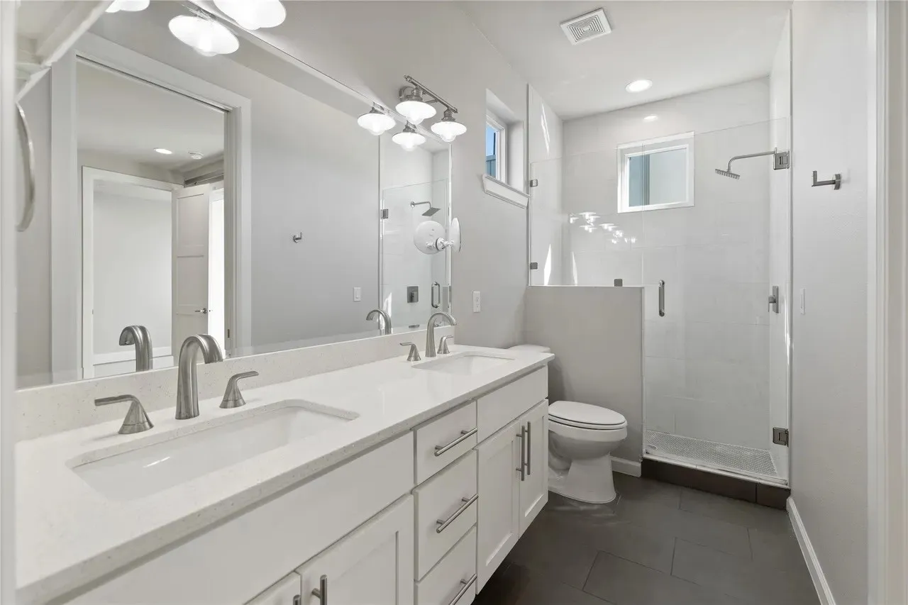Dual vanity with white cabinetry and integrated sinks, a glass-enclosed shower with a rainfall showerhead, large format floor tiles, and a wall-mounted mirror