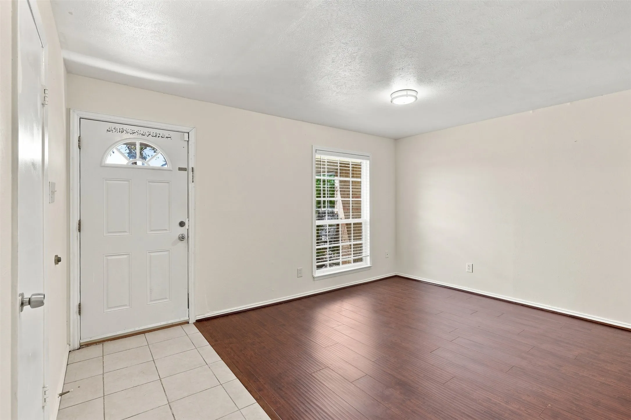 Entryway with tile flooring, leading to a room featuring wood-finish flooring, a window with blinds, and a ceiling-mounted light fixture