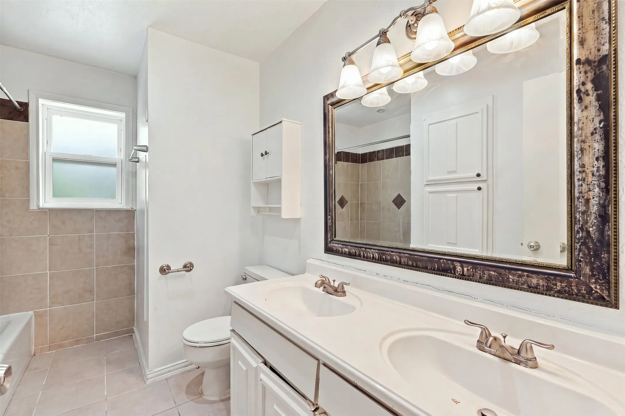 Dual vanity with integrated sinks, framed mirror, built-in storage cabinetry, tiled shower surround with accent tile, and a window for natural light