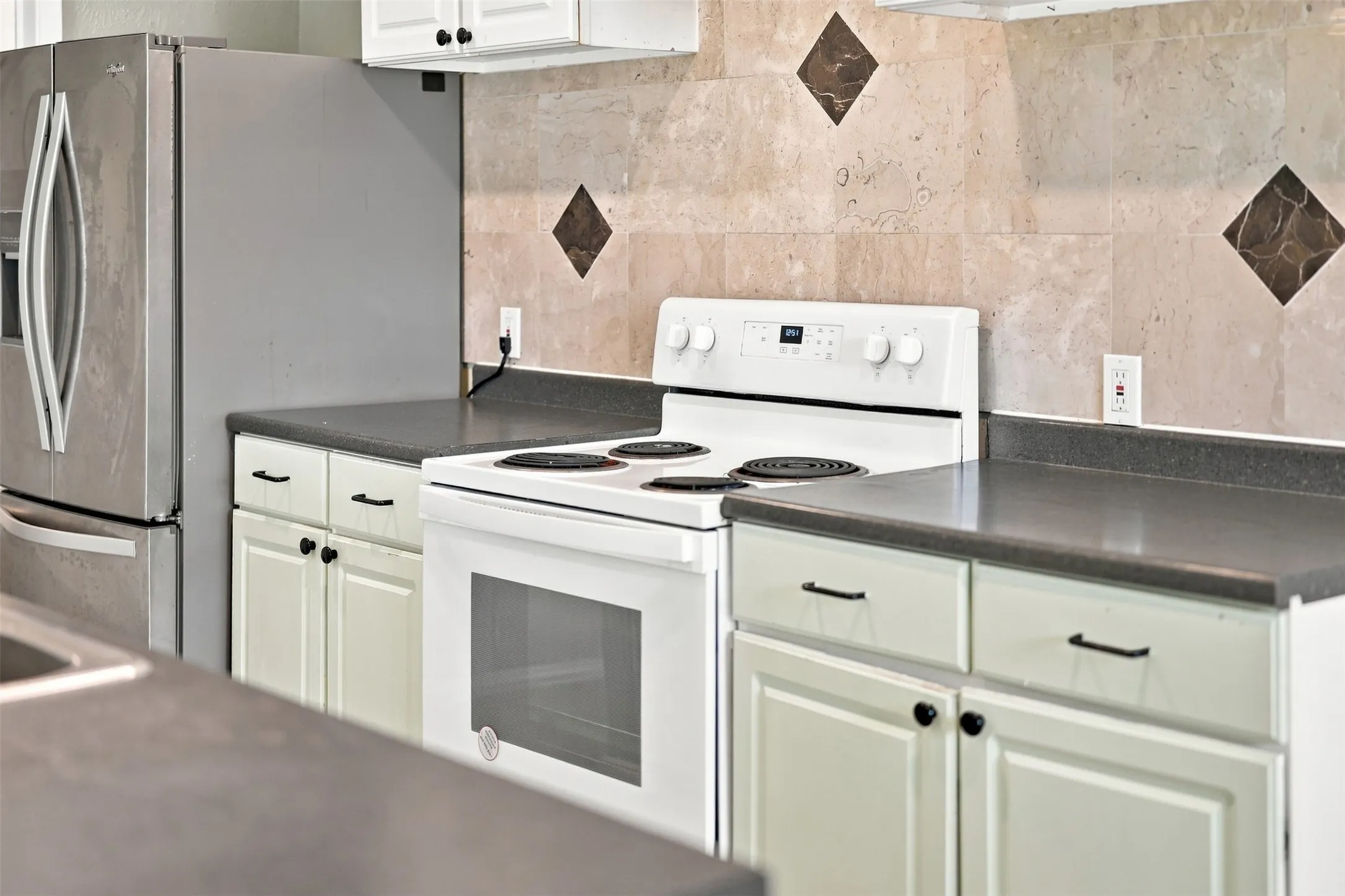 White cabinetry, solid surface countertops, tile backsplash with accent inserts, stainless steel refrigerator, and white freestanding range