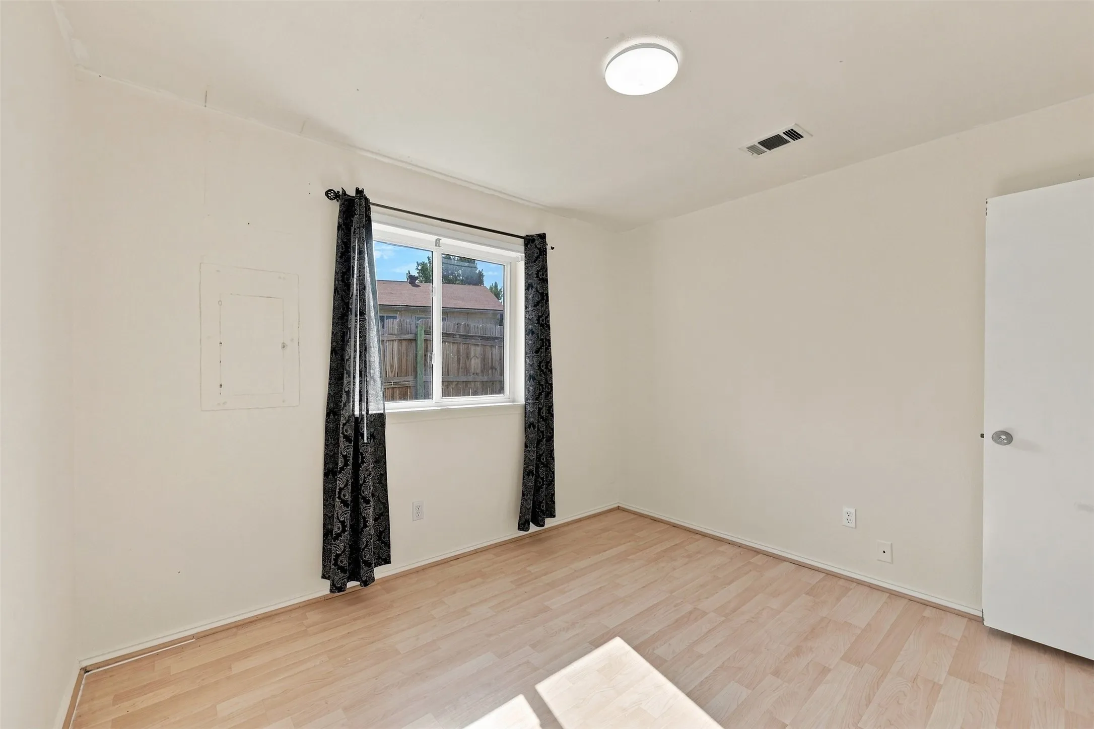 Light wood-finish flooring, white walls, and a single window with black curtain rods