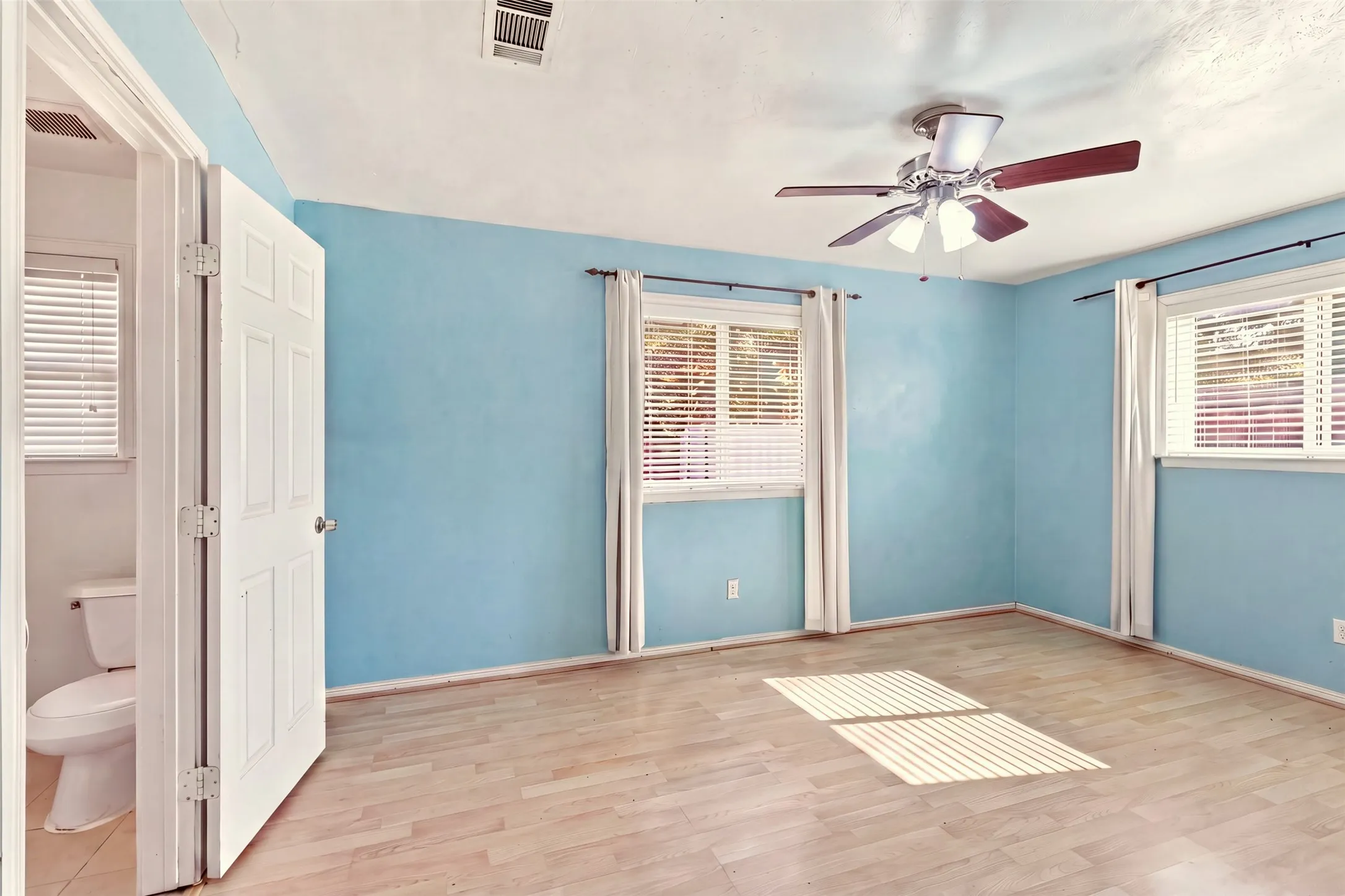Wood-finish flooring, two windows with blinds, ceiling fan with integrated lighting, and direct access to a restroom with a toilet