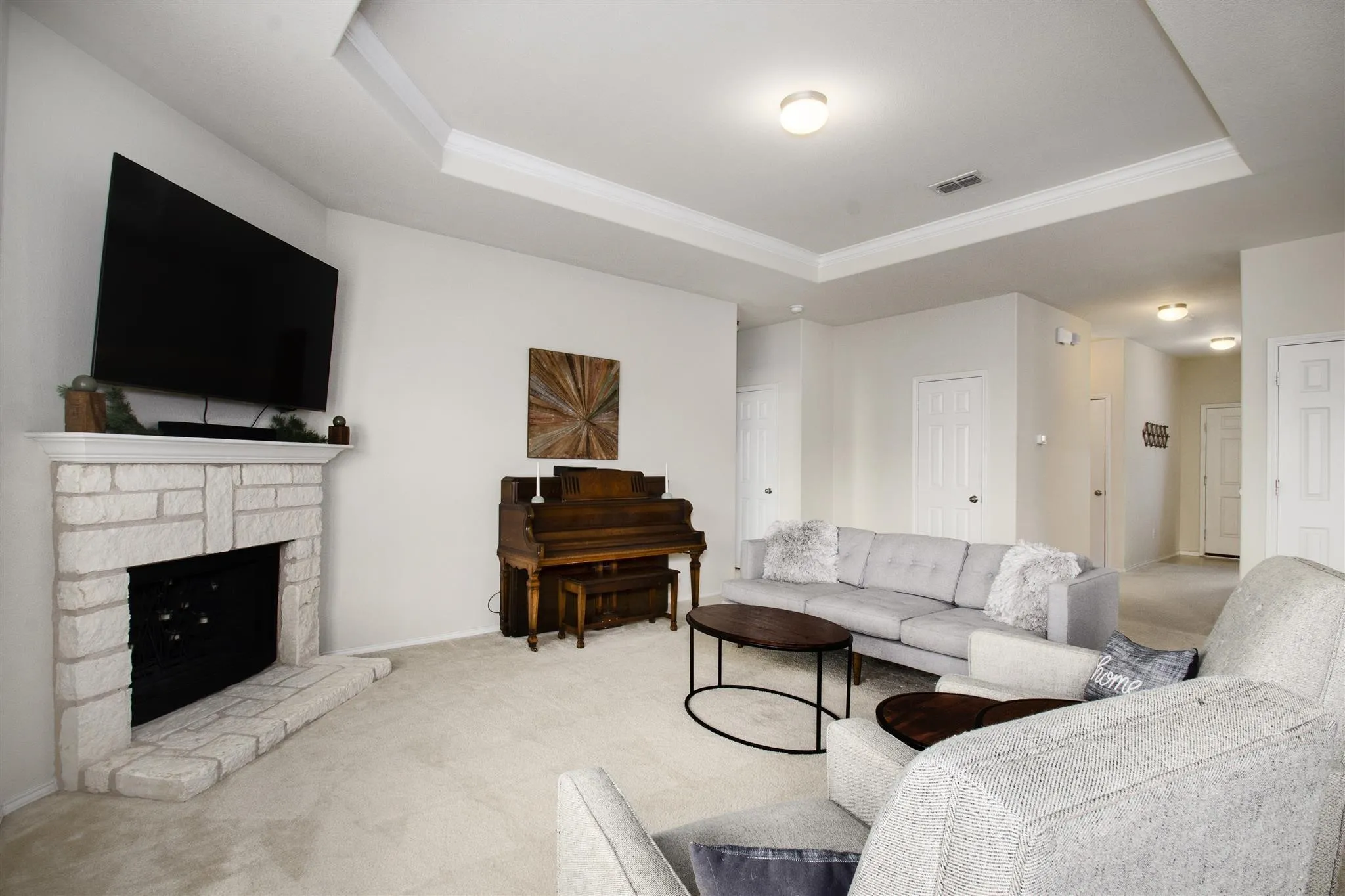 Recessed tray ceiling with crown molding, stone-clad fireplace with mantel, and light-colored carpeting
