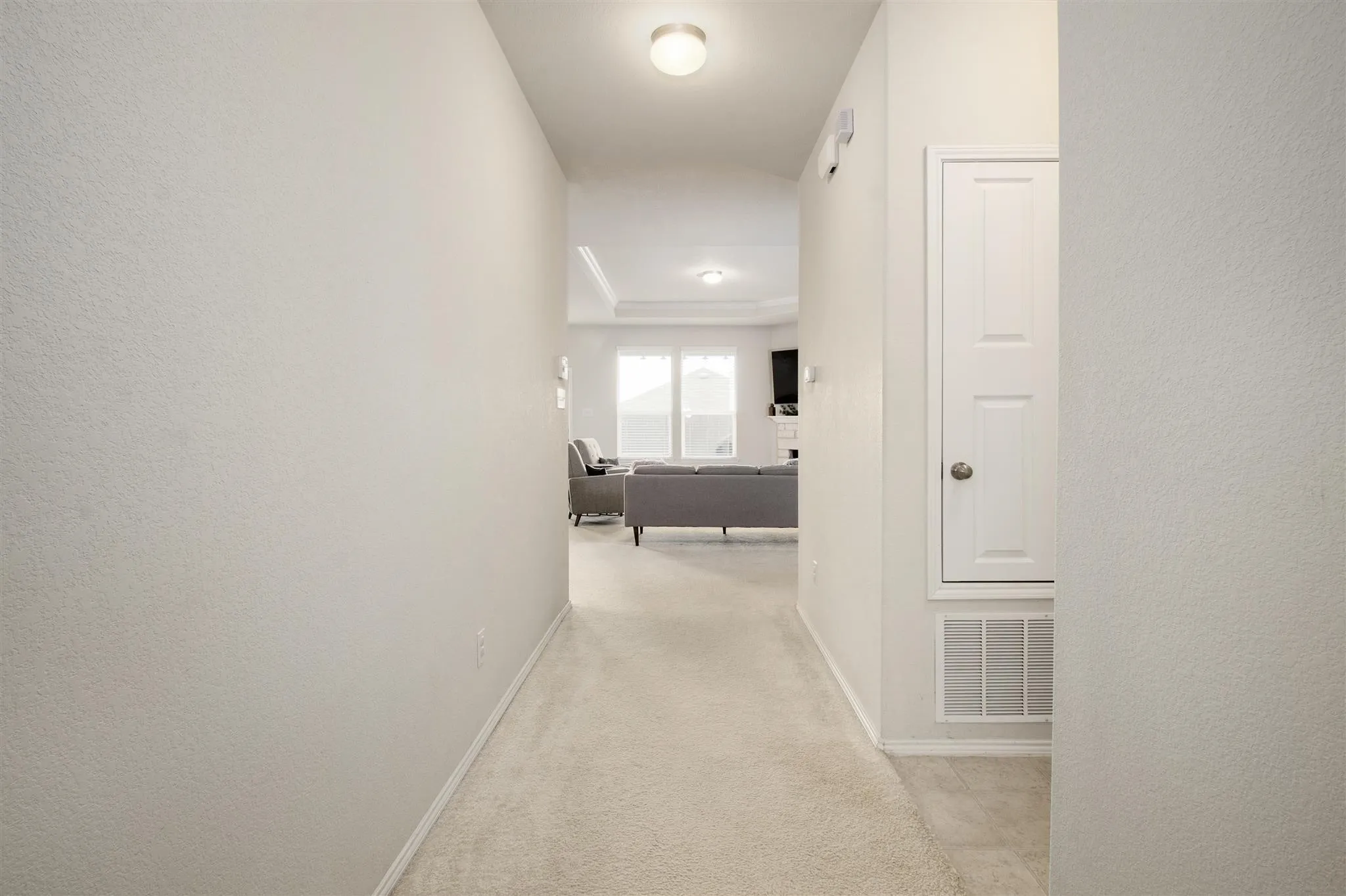 Recessed ceiling lighting illuminates the hallway with plush carpeting, leading to a living area featuring a tray ceiling and bright windows