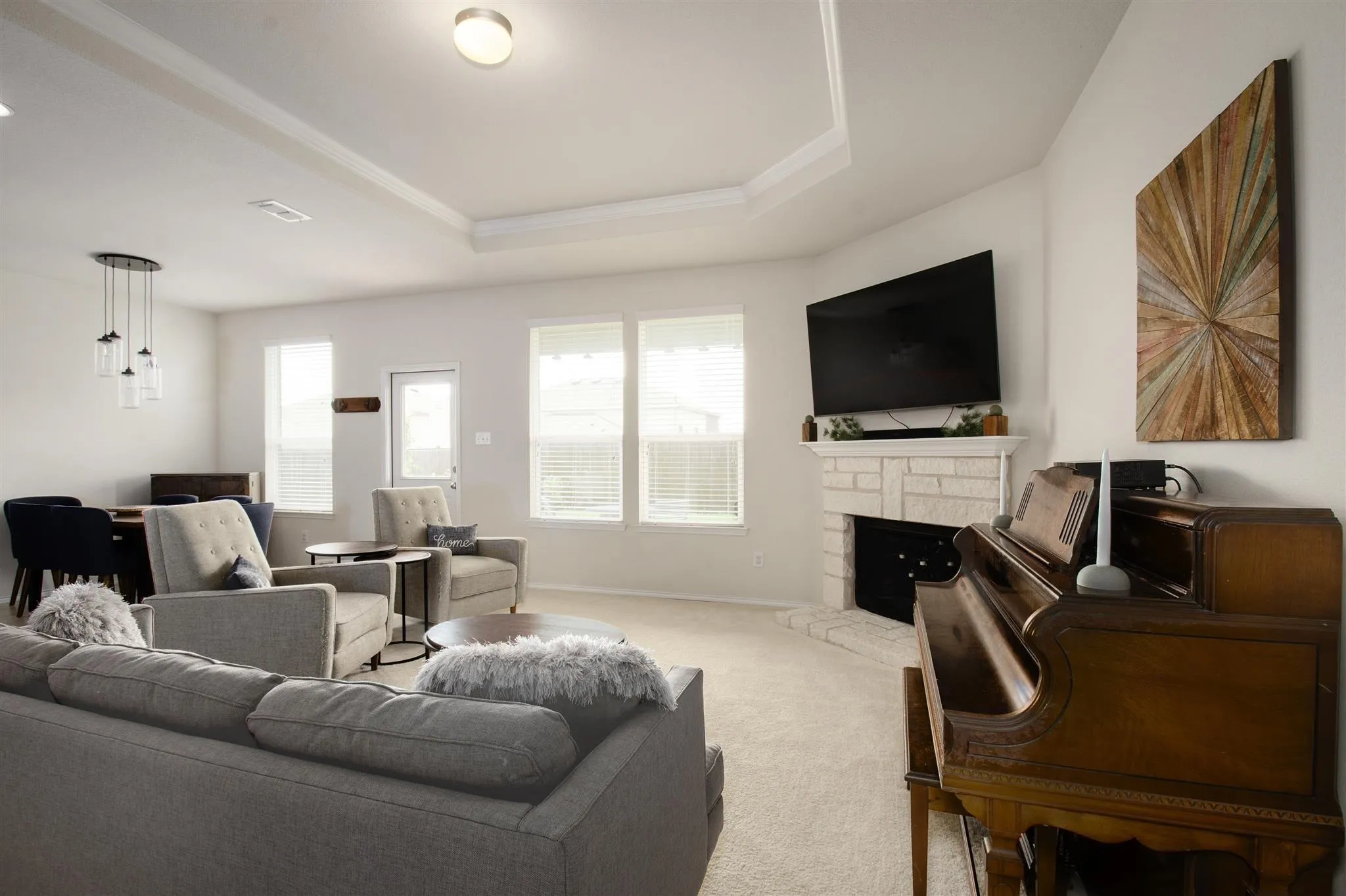 Tray ceiling with crown molding, corner fireplace with stone surround, multiple windows with blinds, built-in ceiling lighting, and a pendant light fixture