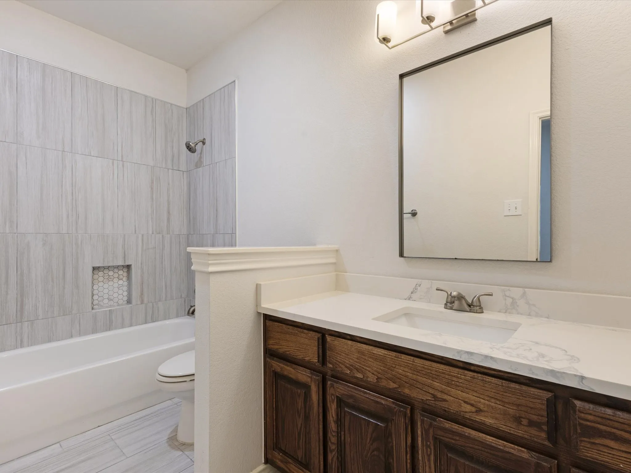 Built-in bathtub with a tile surround and recessed niche, wood-finish vanity with a white countertop and integrated sink, wall-mounted mirror, and overhead light fixture