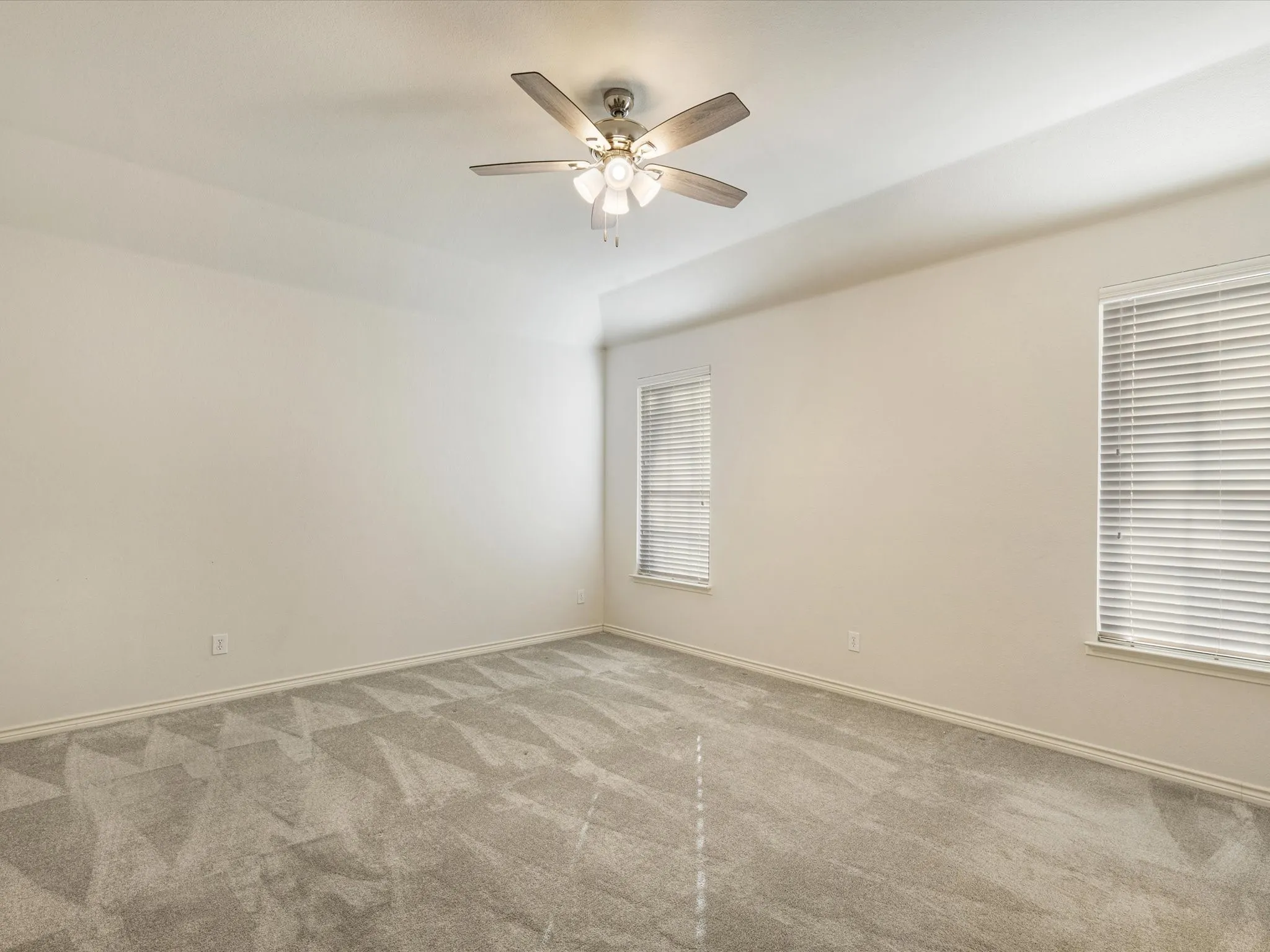Spacious room featuring neutral walls, patterned carpeting, two windows with blinds, a ceiling fan with integrated lighting, and a tray ceiling