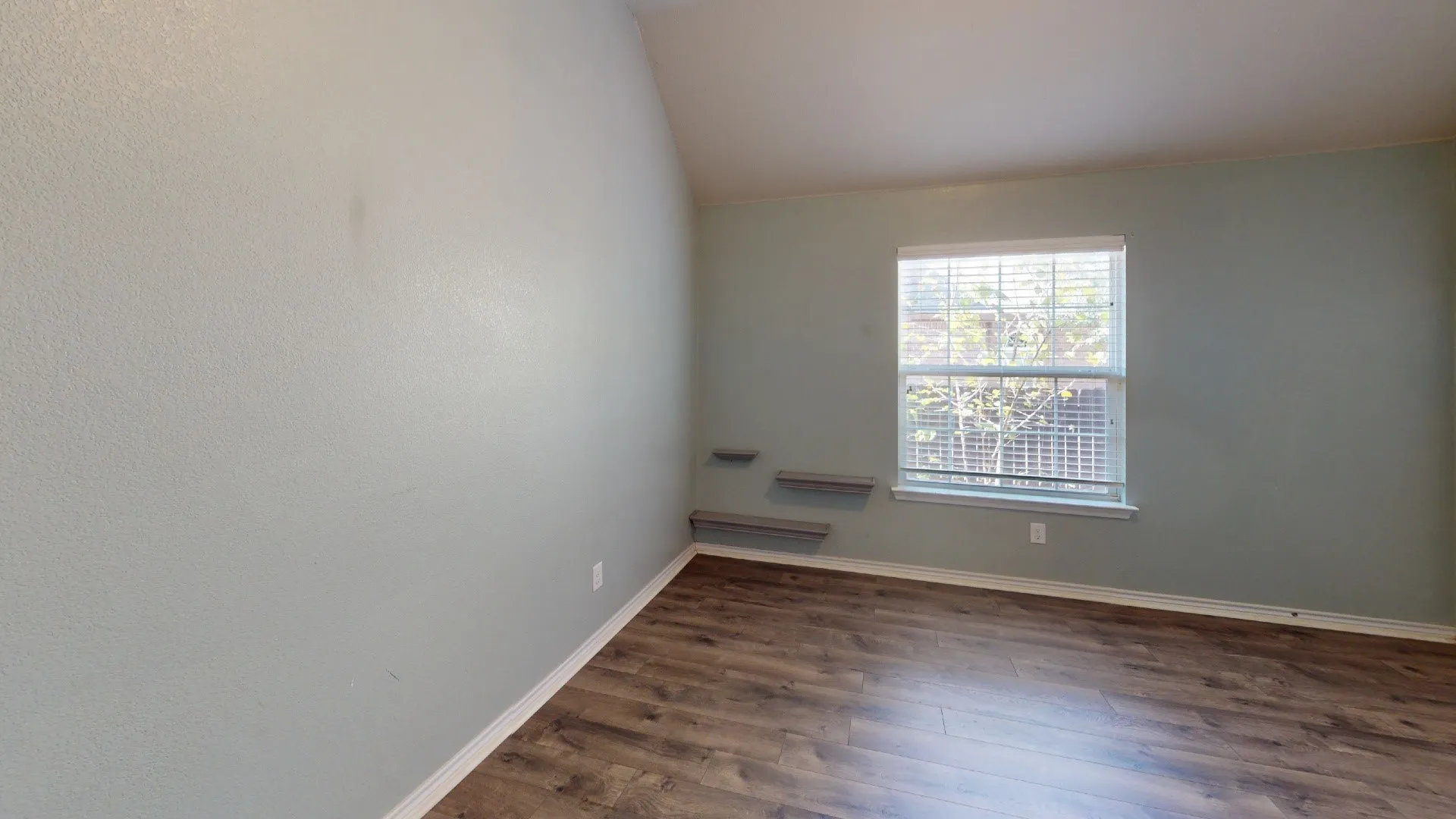 Wood-finish flooring, single window with blinds, light blue wall paint, and two built-in floating shelves