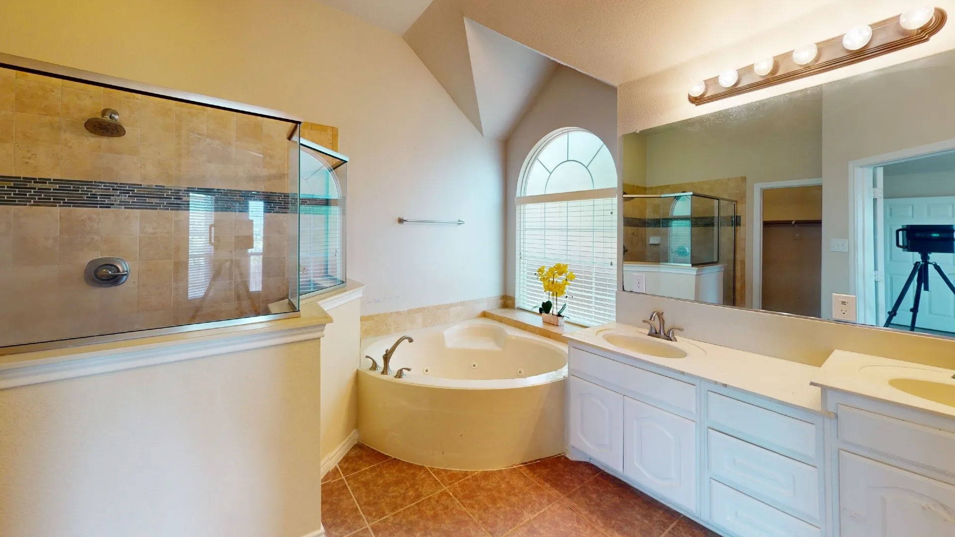 Expansive bathroom featuring a tiled walk-in shower with glass enclosure, a jetted corner tub, a dual vanity with light-toned countertops, and an arched window