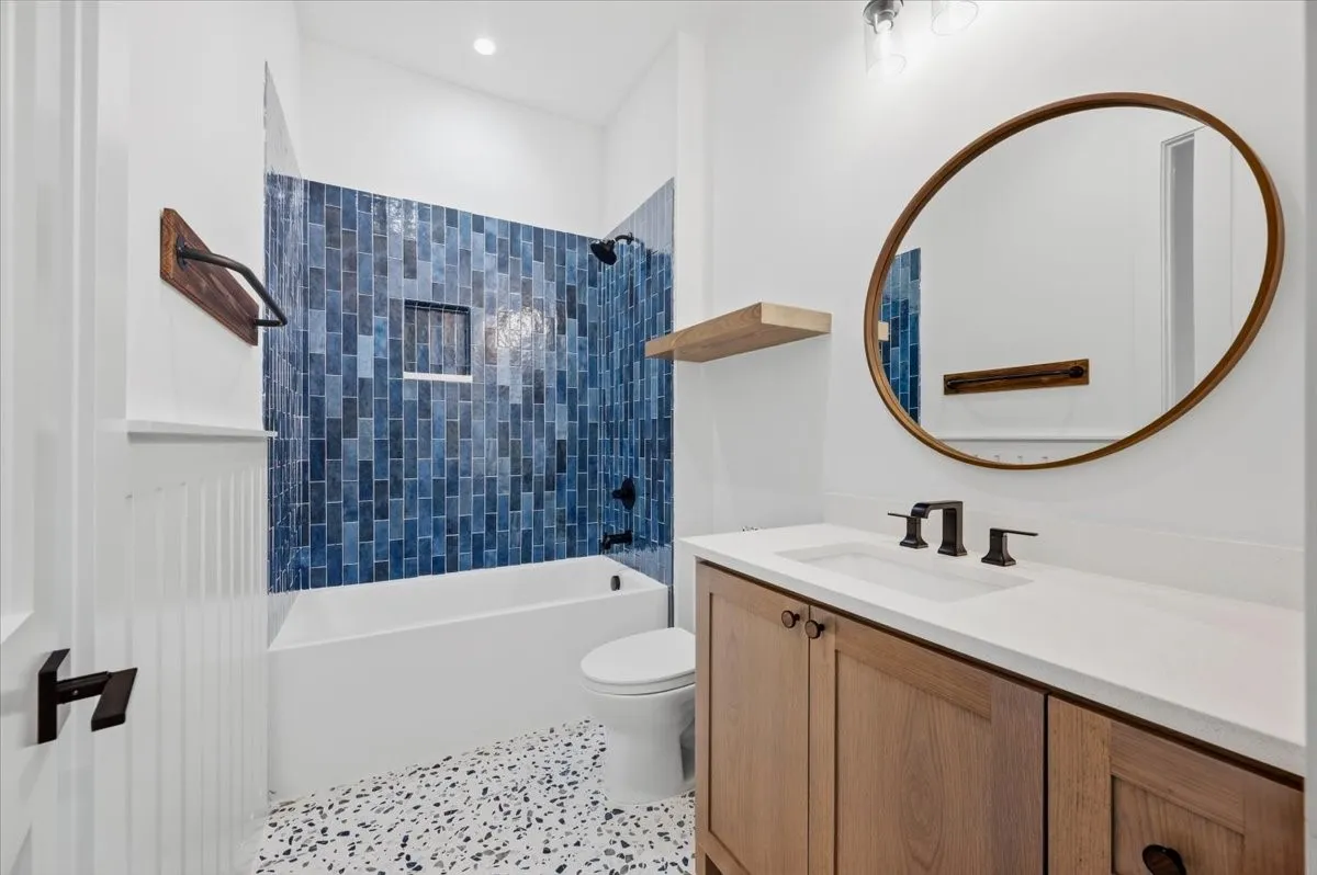 Bathroom featuring blue subway tile shower surround, integrated shower niche, wood-finish vanity with countertop, oval mirror, and speckled flooring