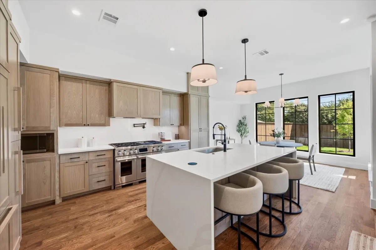 Hardwood flooring throughout, a large kitchen island with counter seating, stainless steel appliances, light wood-finish cabinetry, and black-framed windows