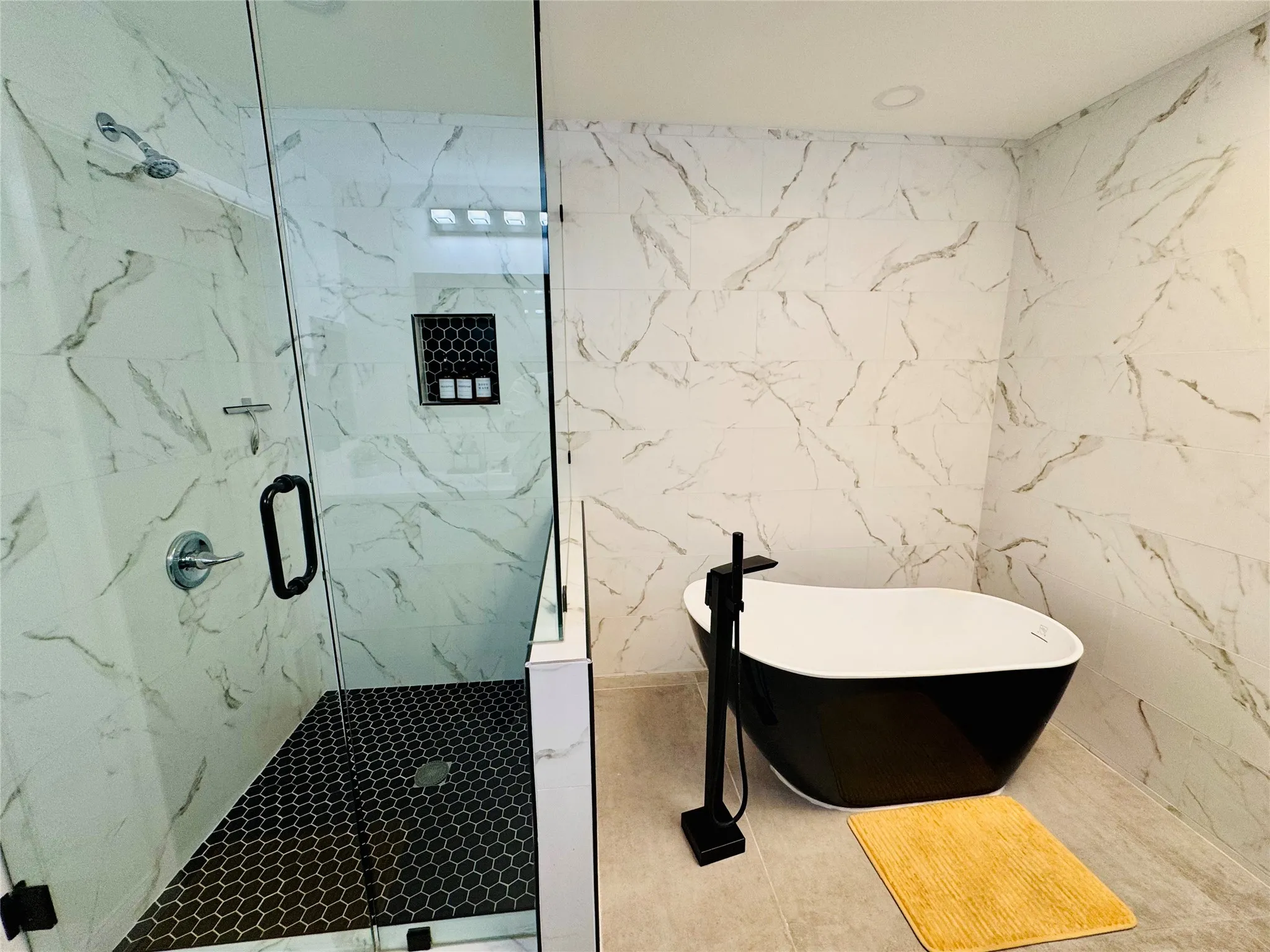 Freestanding soaking tub with a floor-mounted matte black faucet, glass-enclosed shower, black hexagonal tile shower floor, and large format wall tile