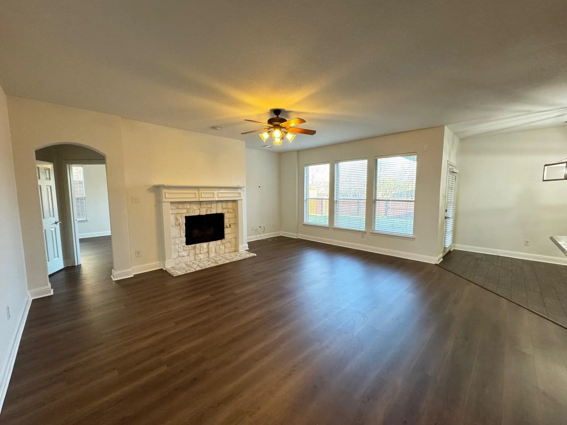 Spacious room featuring wood-finish flooring, a stone-surround fireplace with a white mantel, an arched entryway, three large windows, and a ceiling fan