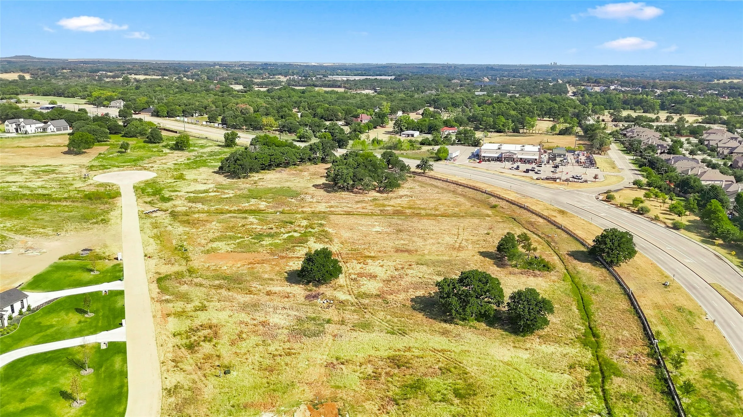 Expansive undeveloped land featuring mature trees, paved roads, and nearby commercial structures