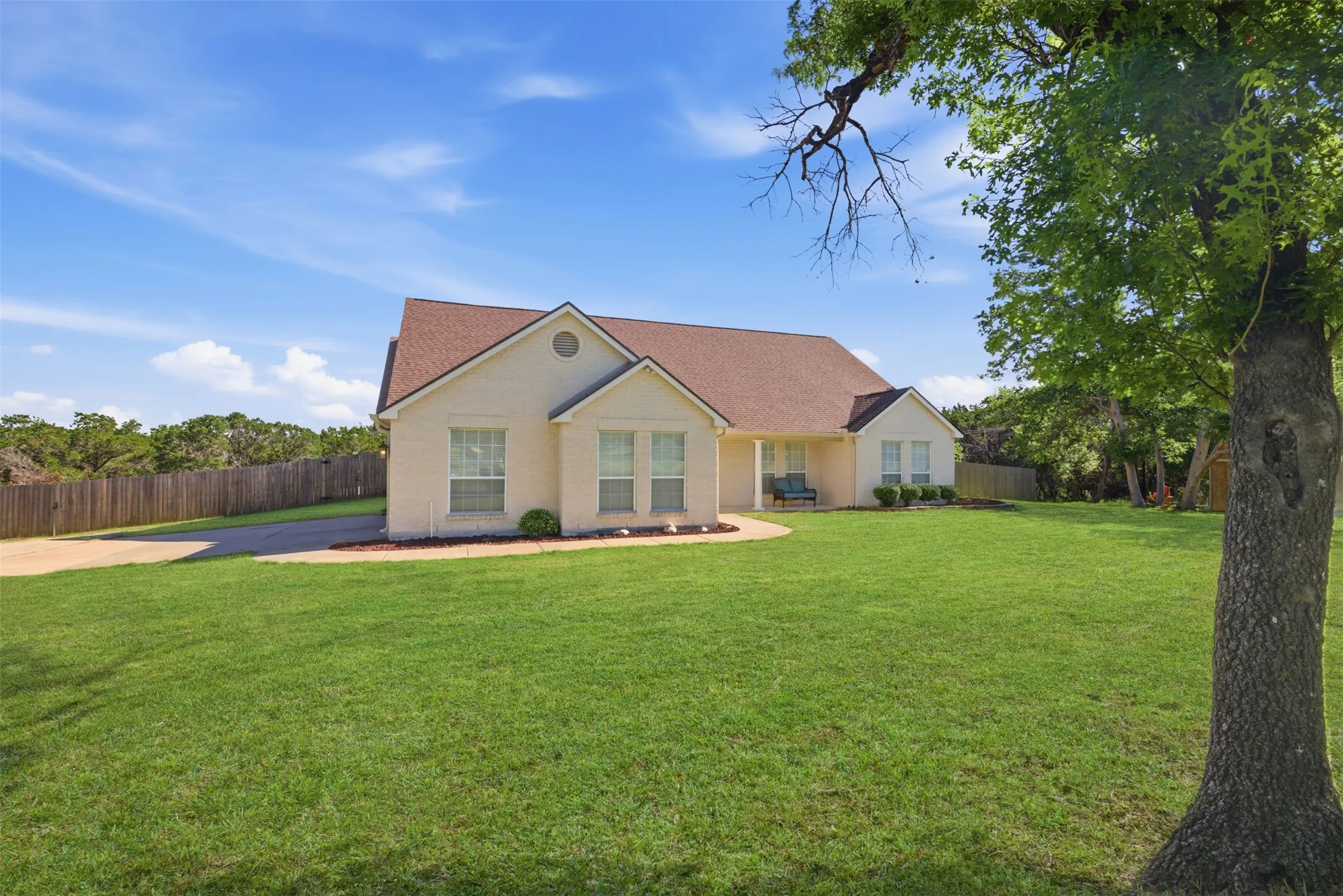 Cream stucco exterior with a brown shingle roof, a paved driveway, and a wooden privacy fence