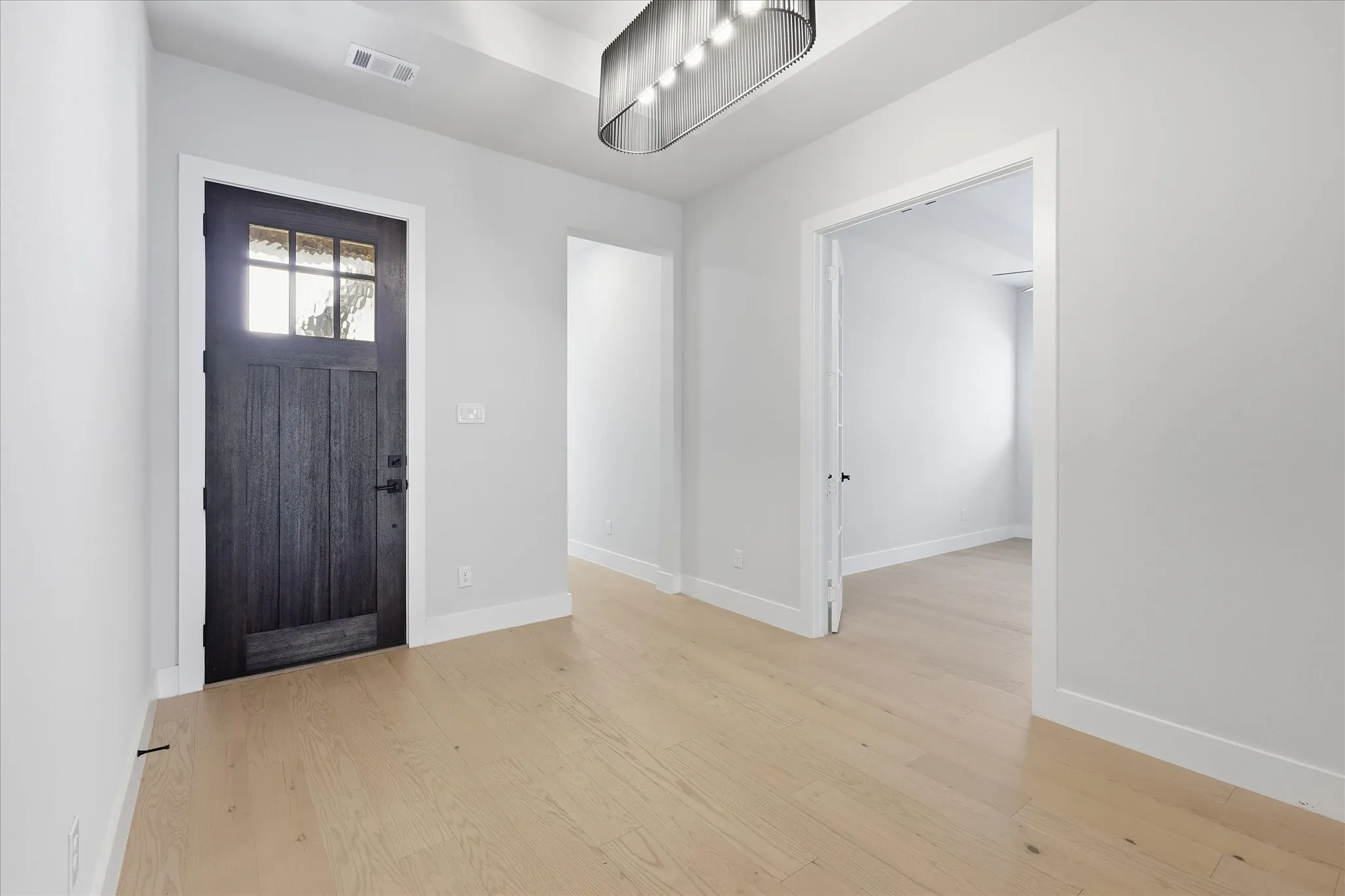 Light wood-finish flooring, dark wood-panel entry door with clear glass, modern linear light fixture, and light gray wall paint