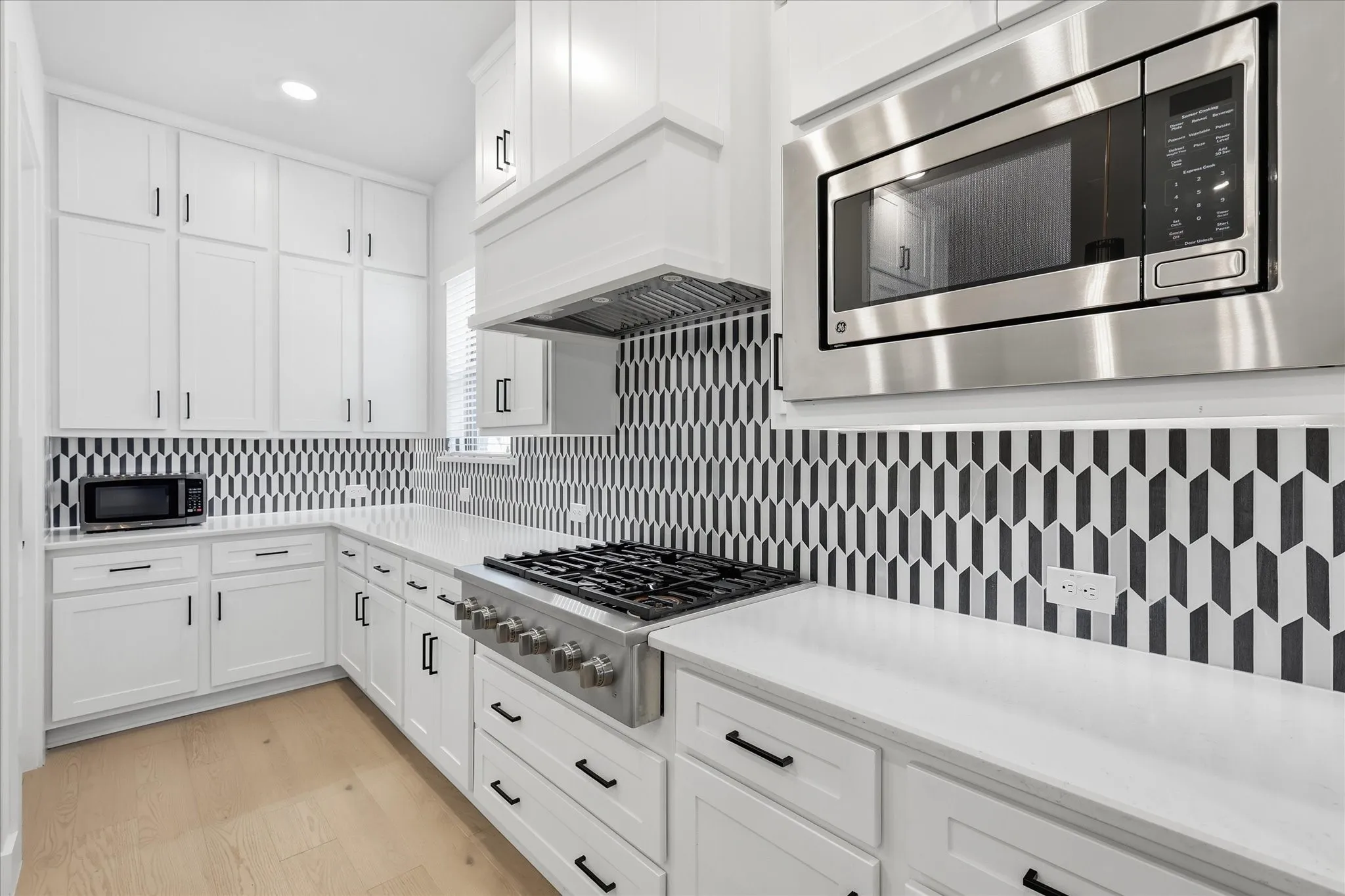 White shaker-style cabinetry, stainless steel appliances, geometric pattern backsplash, light-toned wood-finish flooring, and white countertops