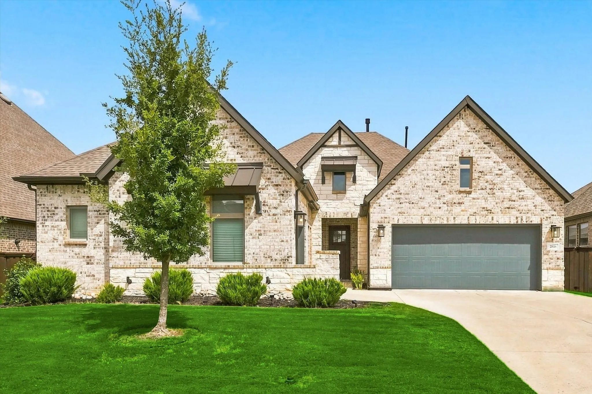 Brick and stone facade with a two-car garage, manicured lawn, and mature tree