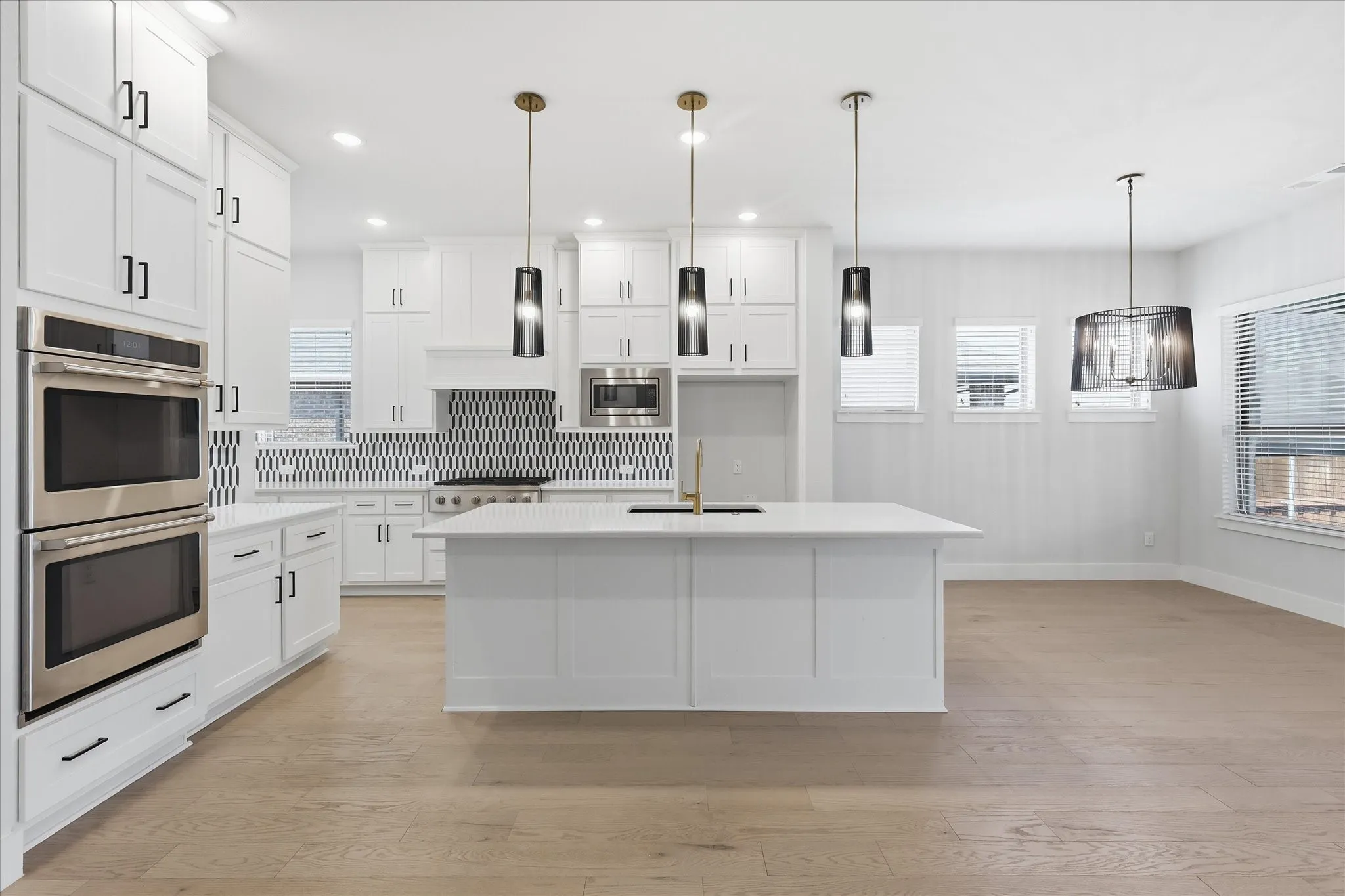 Modern kitchen featuring light wood-finish flooring, white cabinetry with black hardware, stainless steel double ovens, a kitchen island with a built-in sink, and a contrasting black and white tile backsplash