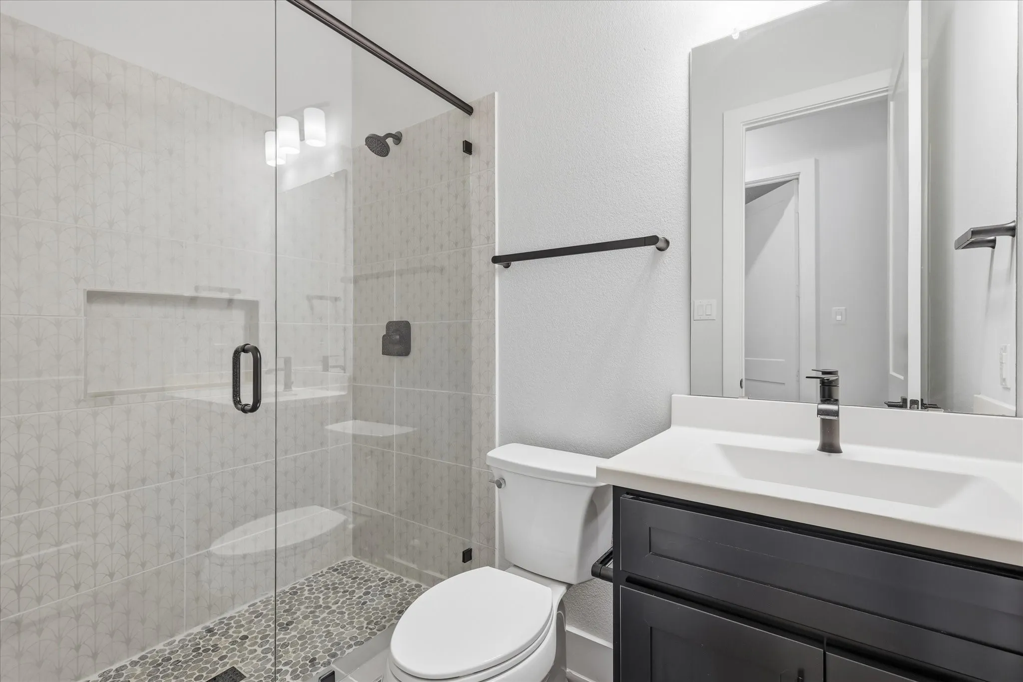 Frameless glass shower enclosure with a recessed shelf, pebble-finish shower pan, and matte black fixtures