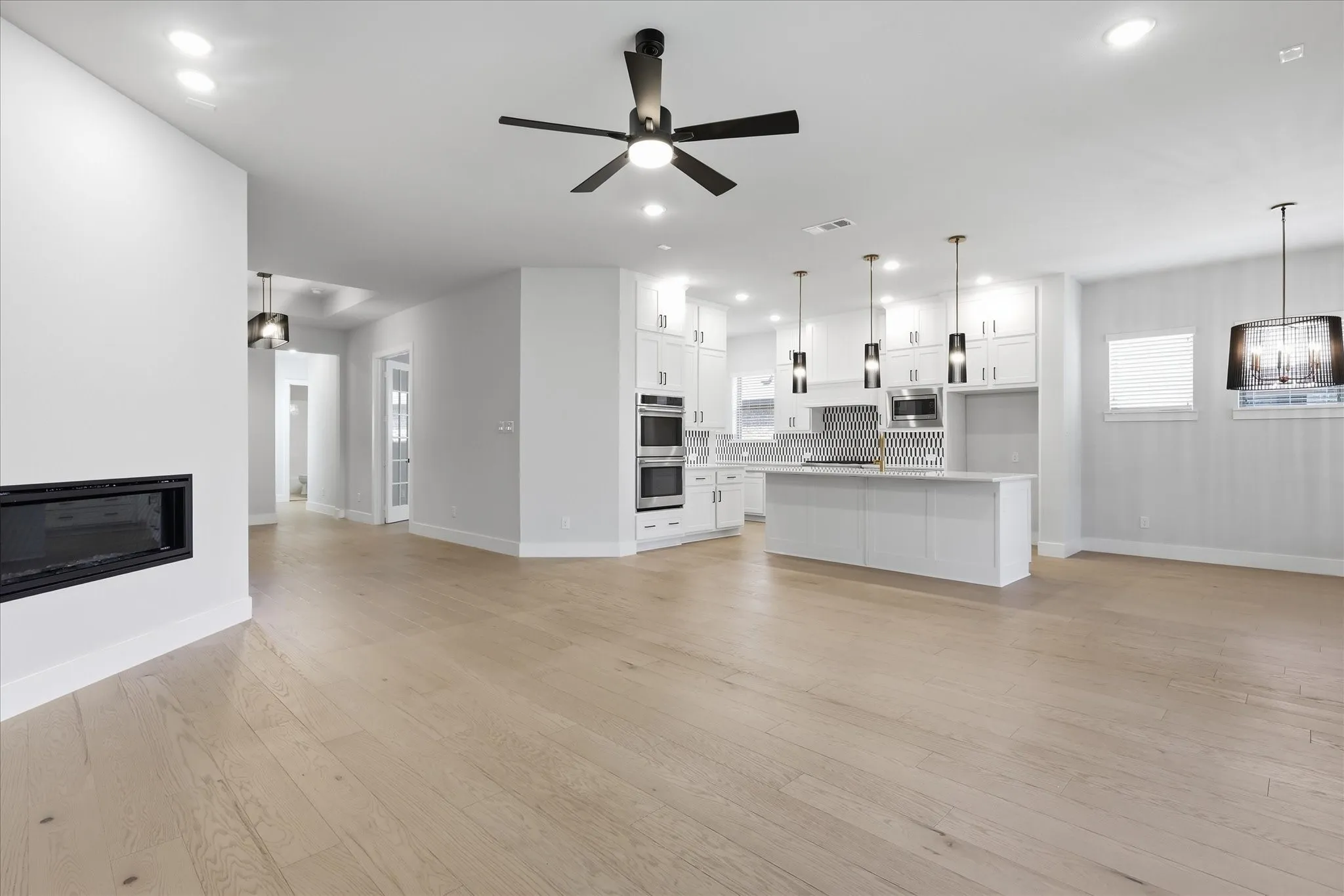 Spacious interior featuring wood-finish flooring, a built-in linear fireplace, a kitchen island with pendant lighting, integrated stainless steel appliances, and white cabinetry