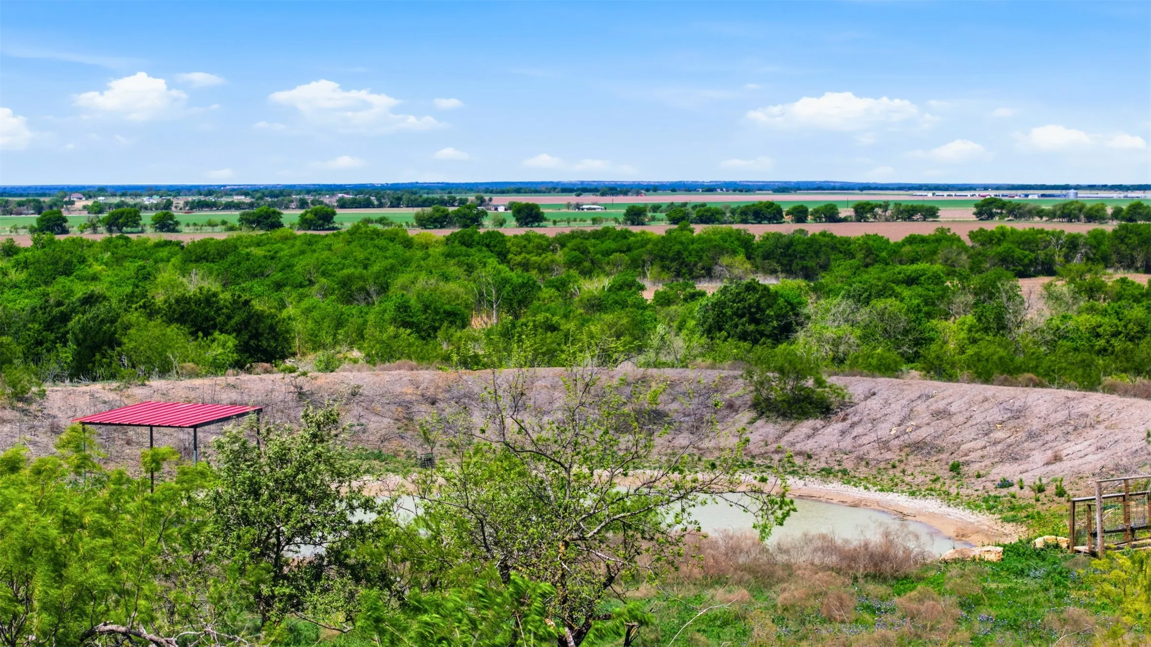 Expansive land featuring a water retention pond, natural foliage, a red-roofed structure, and a distant tree line