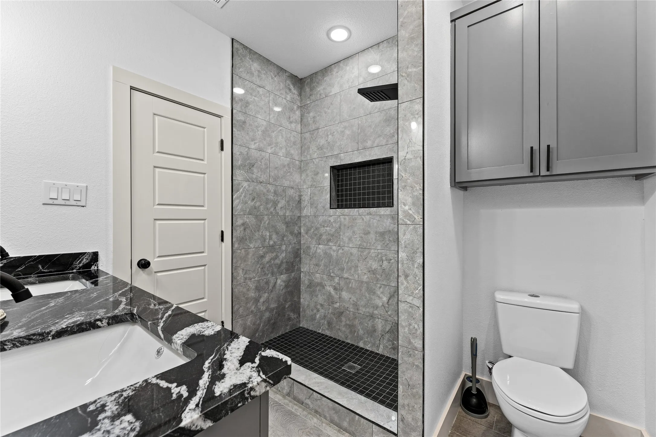 Black and white patterned vanity countertop, undermount sink, walk-in shower with gray tile surround, built-in shower niche, and gray overhead cabinetry
