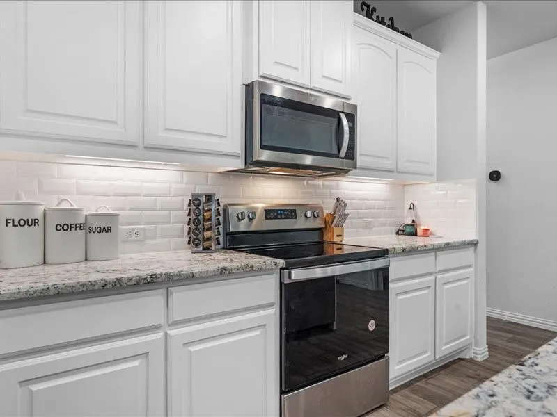 White cabinetry with brushed nickel hardware, granite countertops, subway tile backsplash, stainless steel microwave, and wood-finish flooring