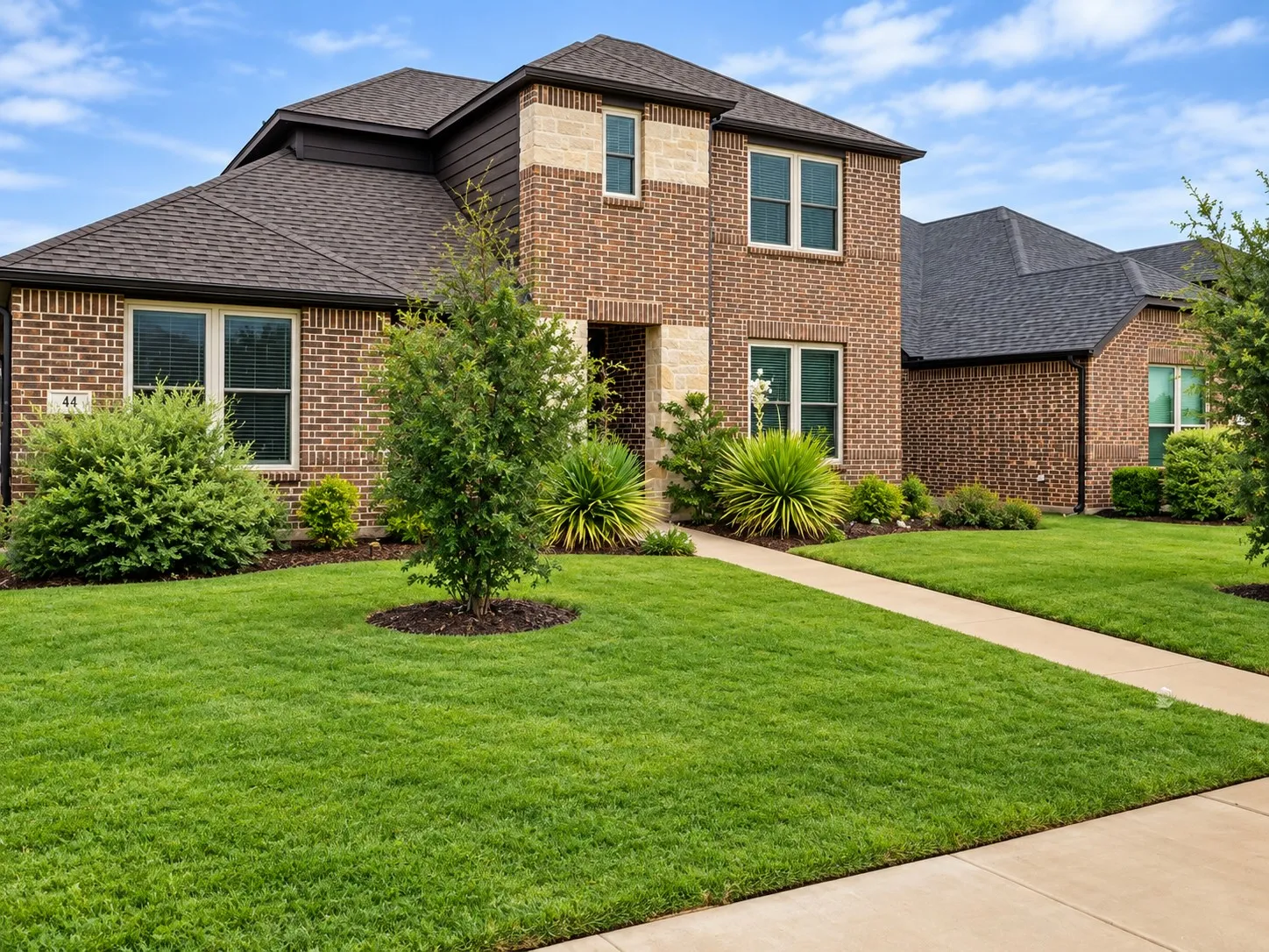 Brick exterior with stone accents, dark-toned roof, front landscaping, mature shrubbery, and a concrete walkway