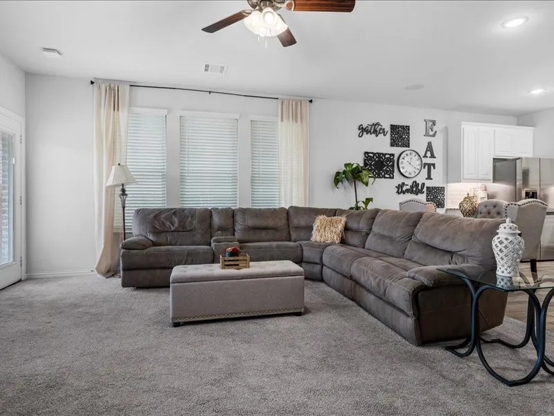 Recessed lighting, ceiling fan, carpet flooring, windows with blinds, and a partial view of a kitchen with white cabinetry