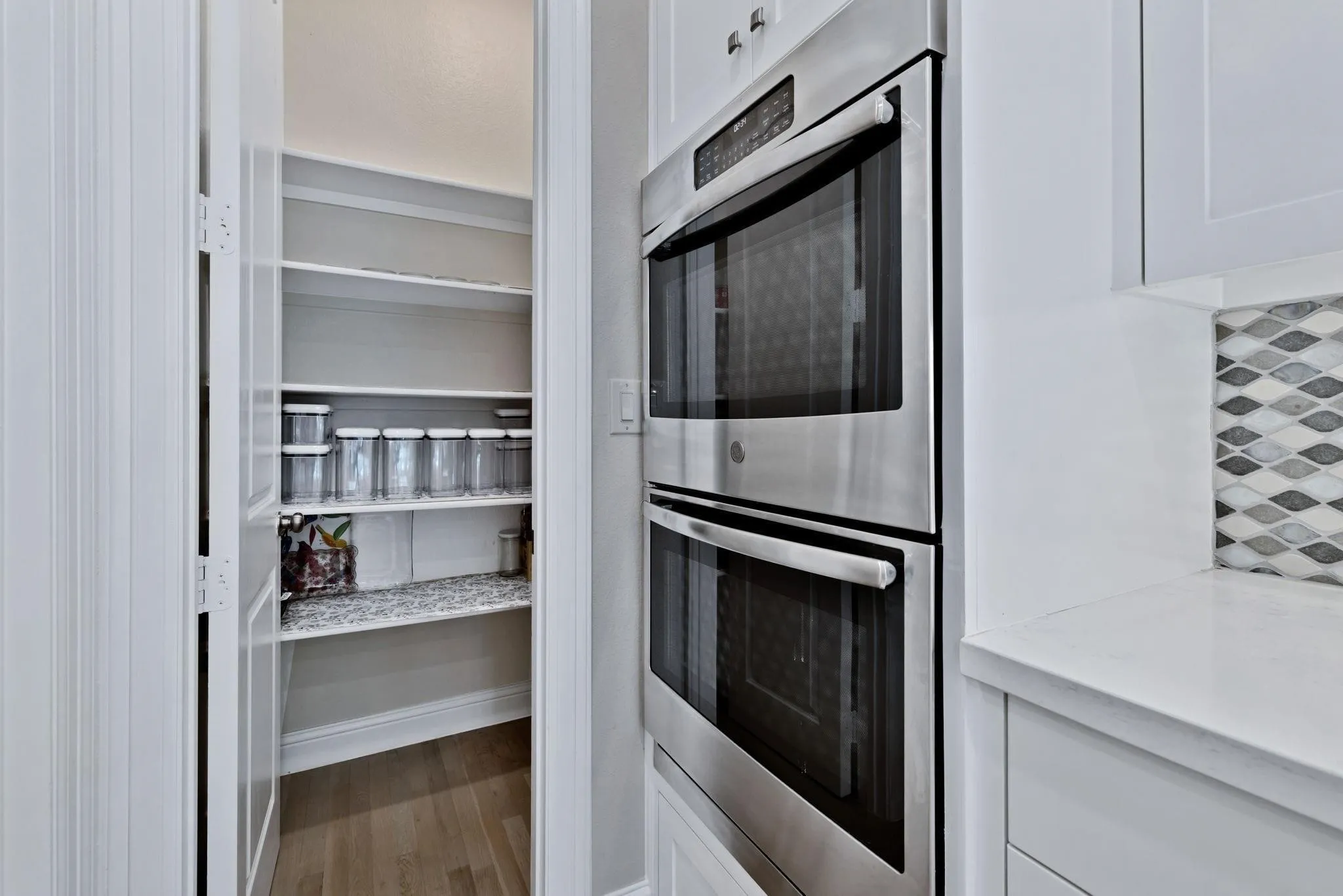 Stainless steel double wall ovens, white cabinetry, white countertop, geometric tile backsplash, and a walk-in pantry with wood-finish flooring