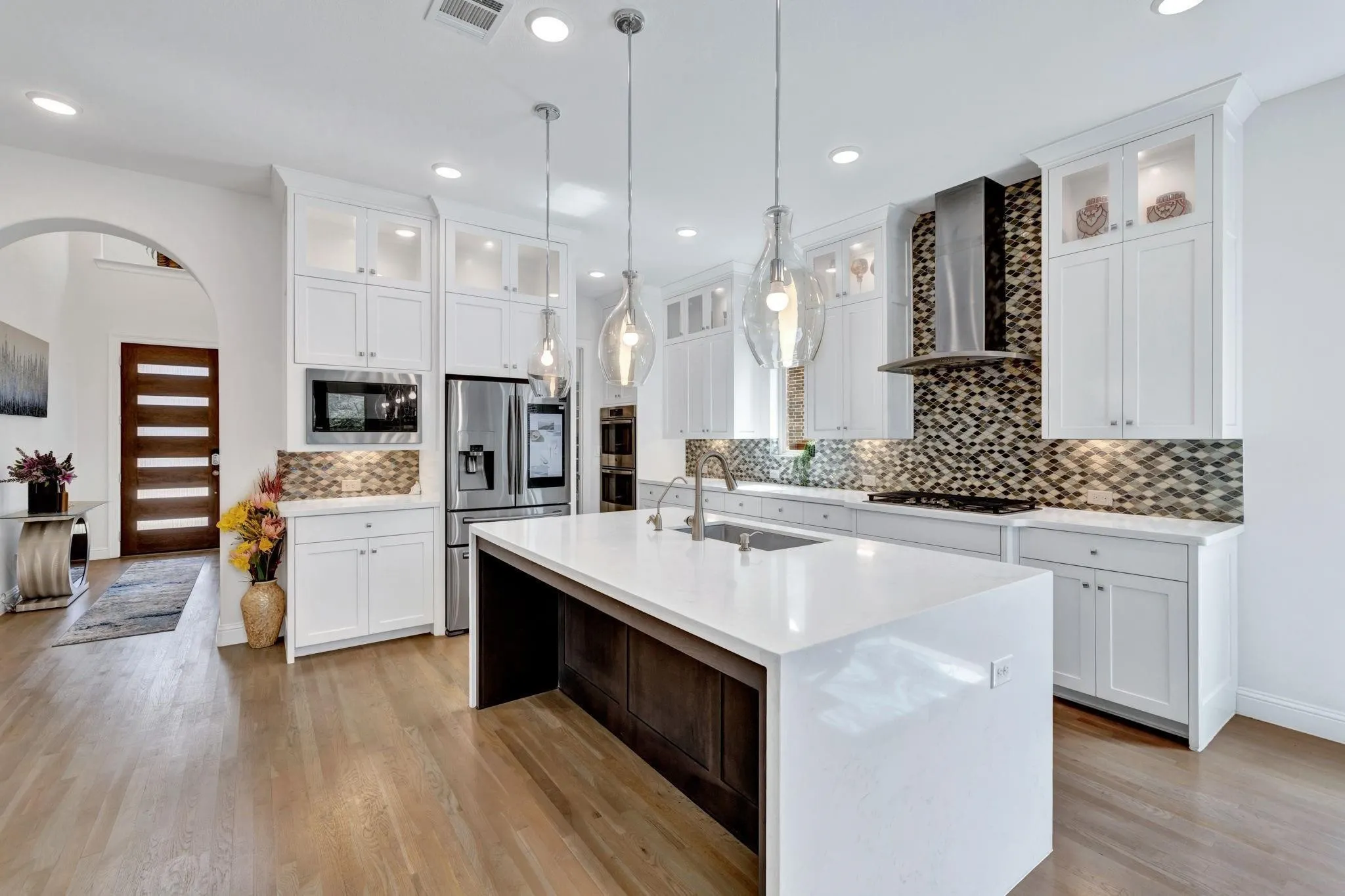 Wood-finish flooring throughout, white cabinetry, stainless steel appliances, mosaic tile backsplash, and a large central island