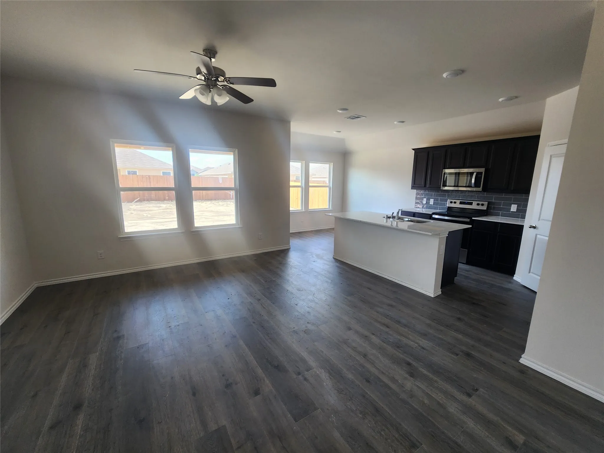 Spacious open-concept layout featuring wood-finish flooring, a kitchen island with a built-in sink, dark cabinetry, and a subway tile backsplash