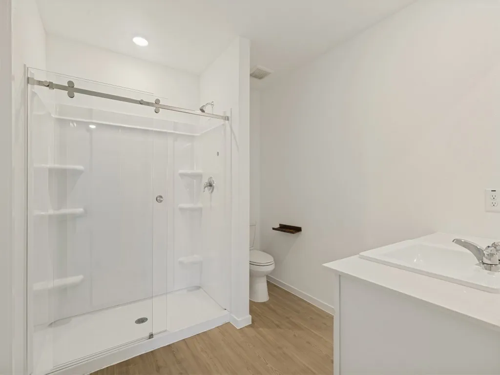 Frameless glass shower enclosure with sliding door, integrated shower shelving, single vanity with integrated sink, chrome faucet, and wood-finish flooring