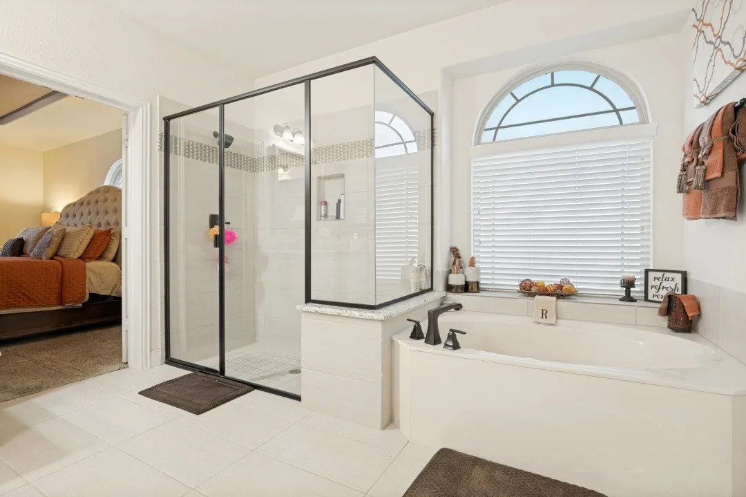 Large soaking tub with dark bronze fixtures, an arched window with blinds, and an enclosed glass shower with dark bronze framing