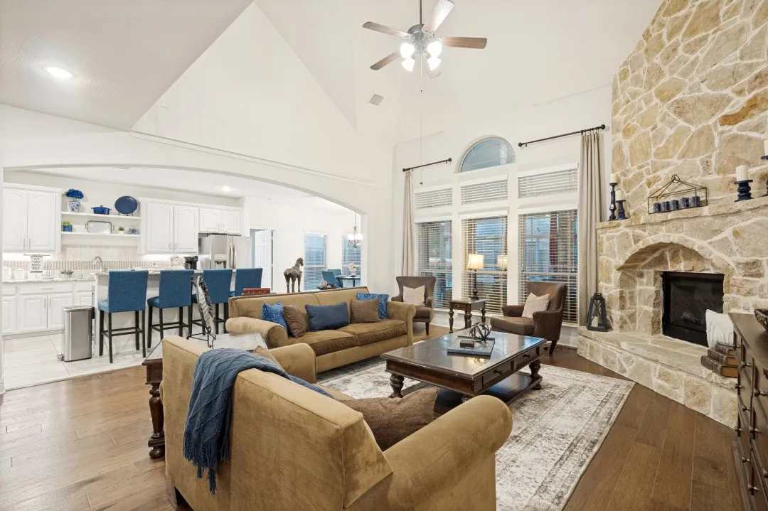 Vaulted ceiling, stone fireplace, arched window, wood-finish flooring, and a kitchen featuring white cabinetry