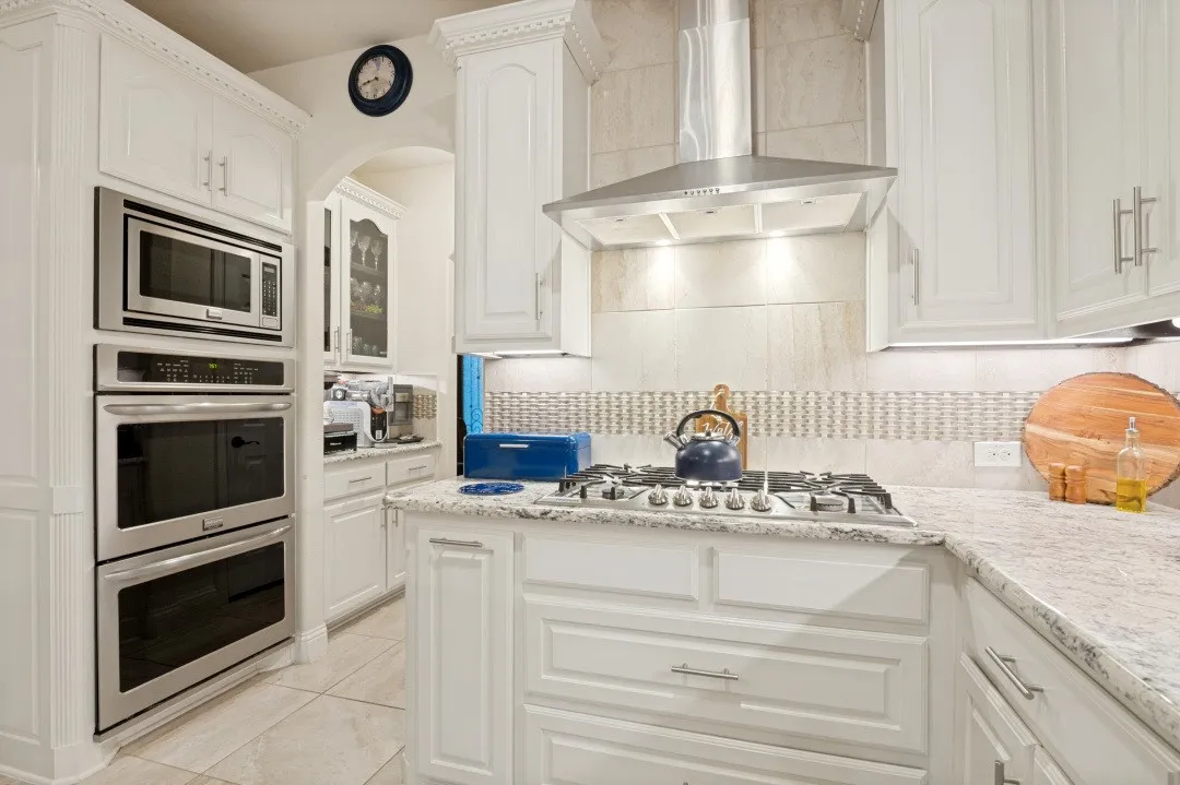 Sophisticated chef's kitchen featuring custom white cabinetry, stainless steel appliances including a gas cooktop with professional-grade range hood, dual wall-mounted ovens, and quartz countertops