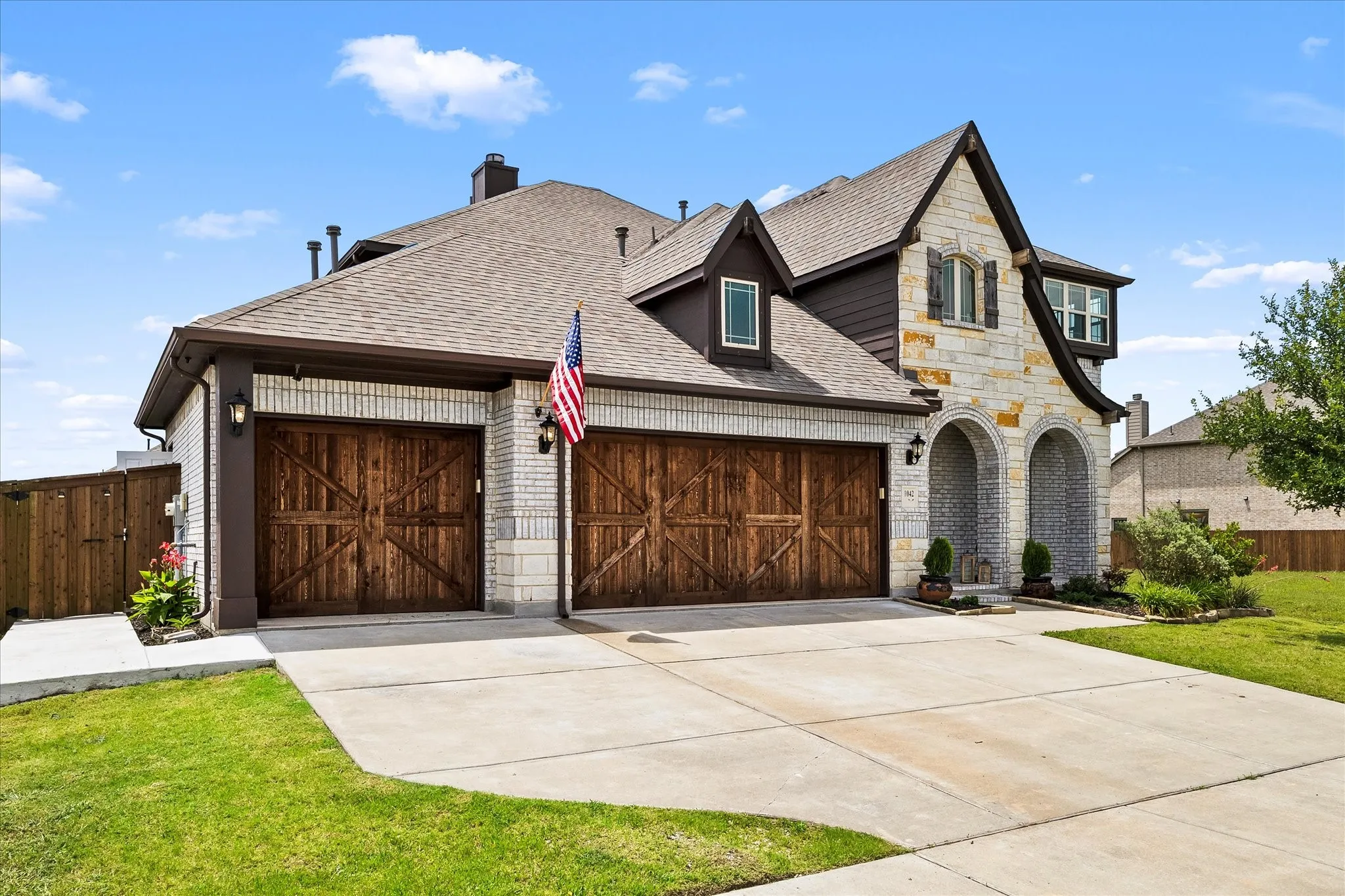 Stone and brick exterior featuring a two-car garage with wood-finish doors, arched entryways, a dormer window, and a gabled roofline