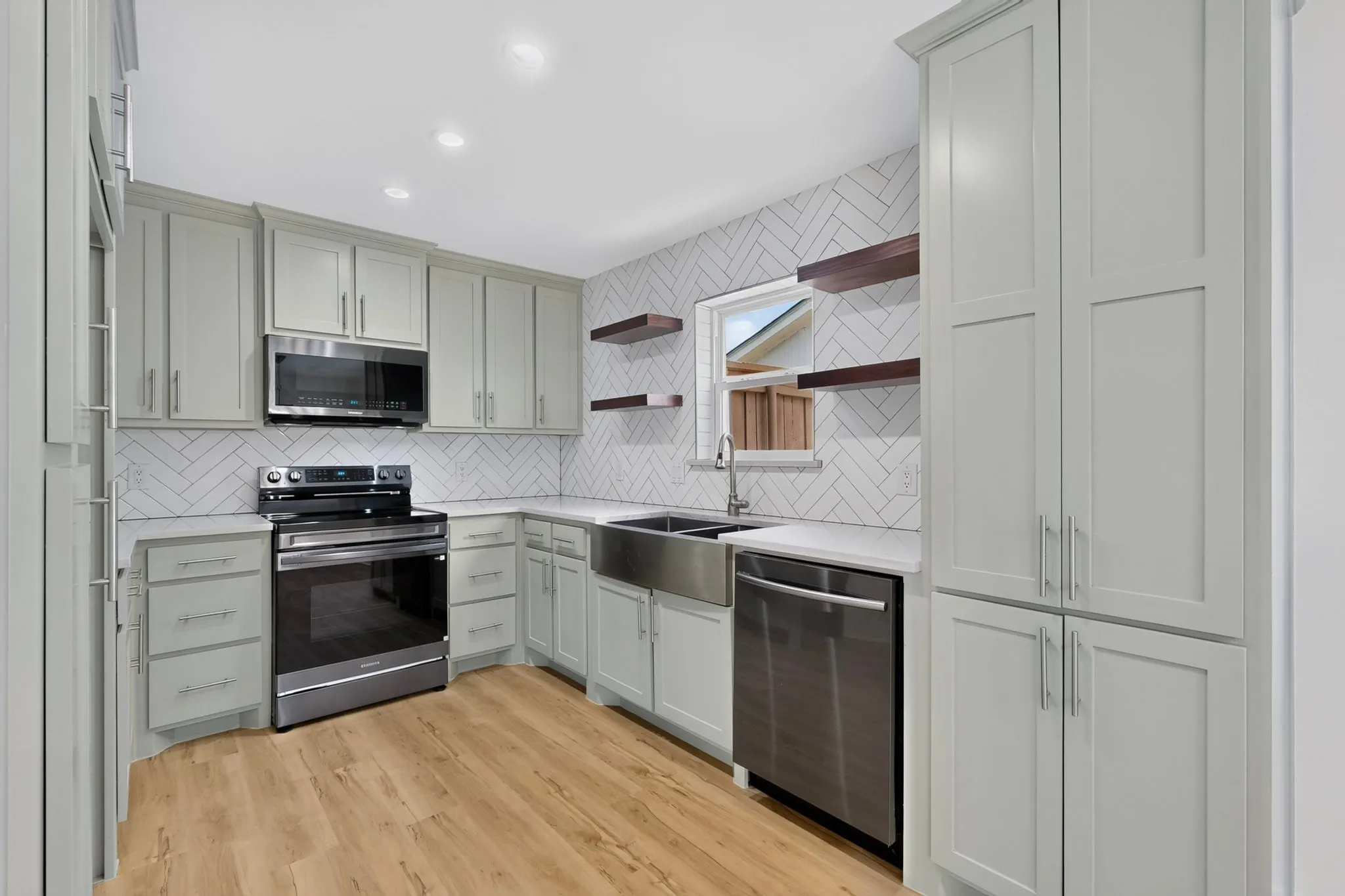 Kitchen featuring light gray cabinetry, stainless steel appliances, wood-finish flooring, white herringbone tile backsplash, and a stainless steel farmhouse sink