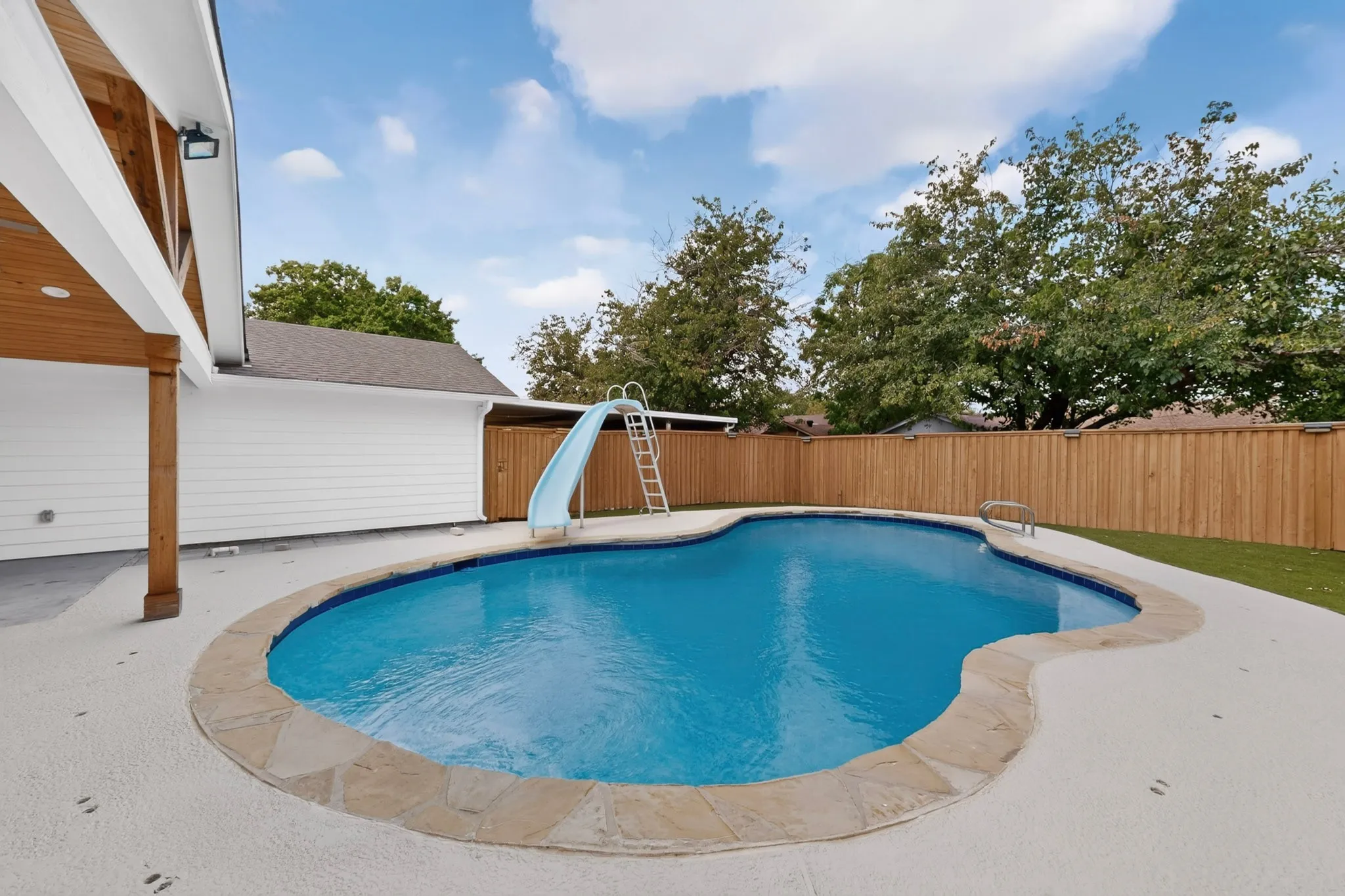 Freeform swimming pool with stone coping, a surrounding concrete deck, and a wooden privacy fence