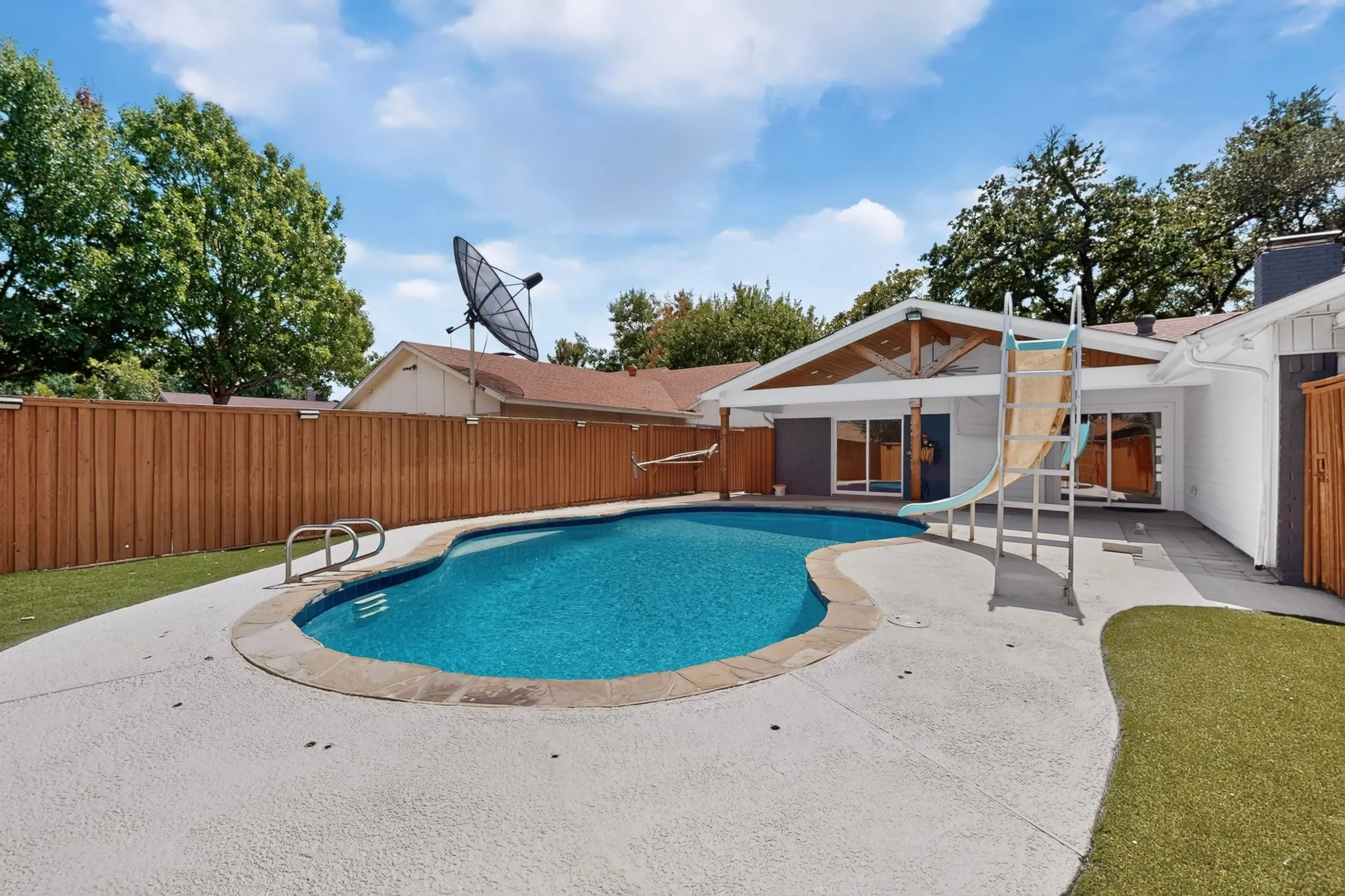Freeform swimming pool with integrated slide, expansive concrete patio, and a wood privacy fence