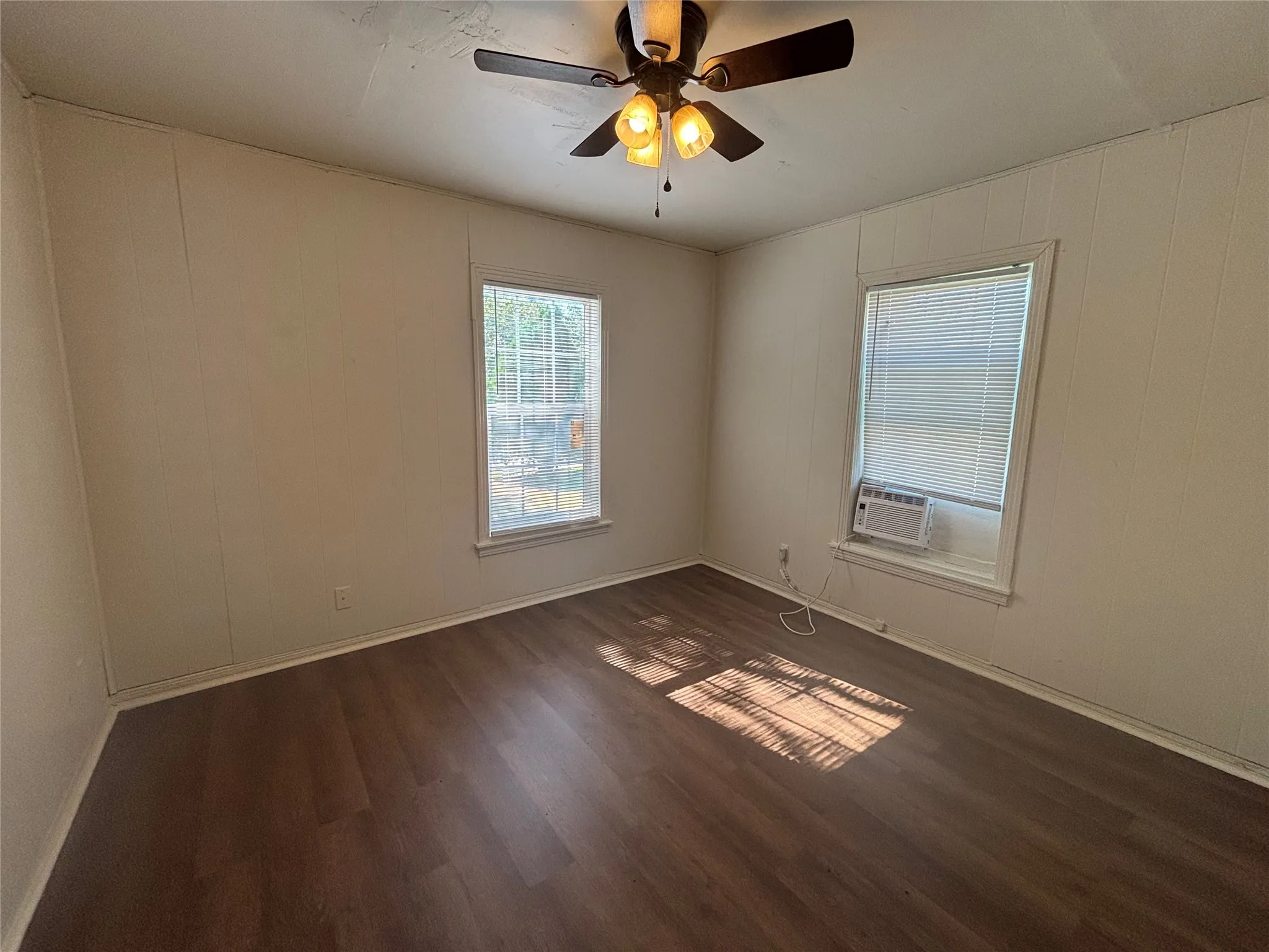 Wood-finish flooring, two windows with blinds, ceiling fan with integrated lighting, and a wall-mounted air conditioning unit