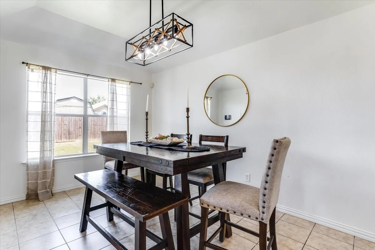 Dining room showcasing a black wood table with bench seating, upholstered side chair with nailhead trim, tile flooring, large round mirror with brass frame, geometric pendant lighting fixture, and abundant natural light from multi-pane windows with sheer curtains