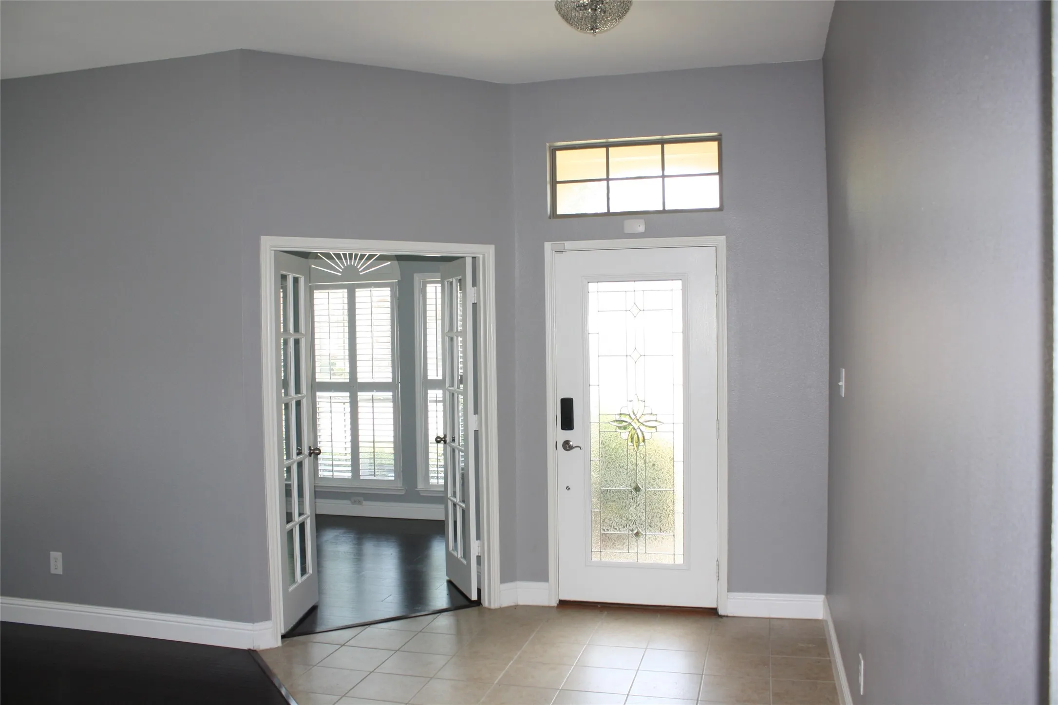 Vaulted ceiling entry with a decorative semi-flush mount fixture, featuring a transom window above the glass-paneled front door, and French doors with decorative pediment leading to a room with wood-finish flooring