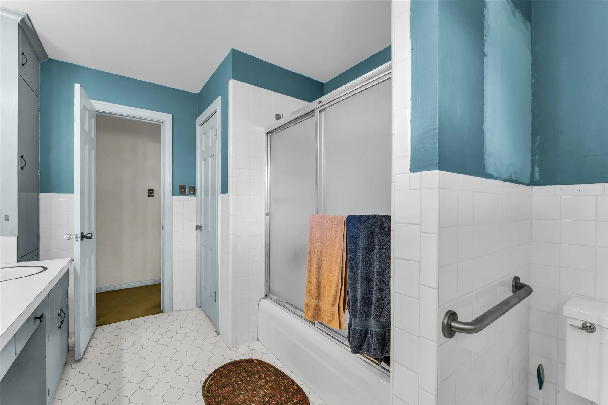 Upstairs; White hexagonal tile flooring, white tile wainscoting, built-in vanity with integrated sink, built-in storage cabinetry, and a bathtub with a sliding glass enclosure