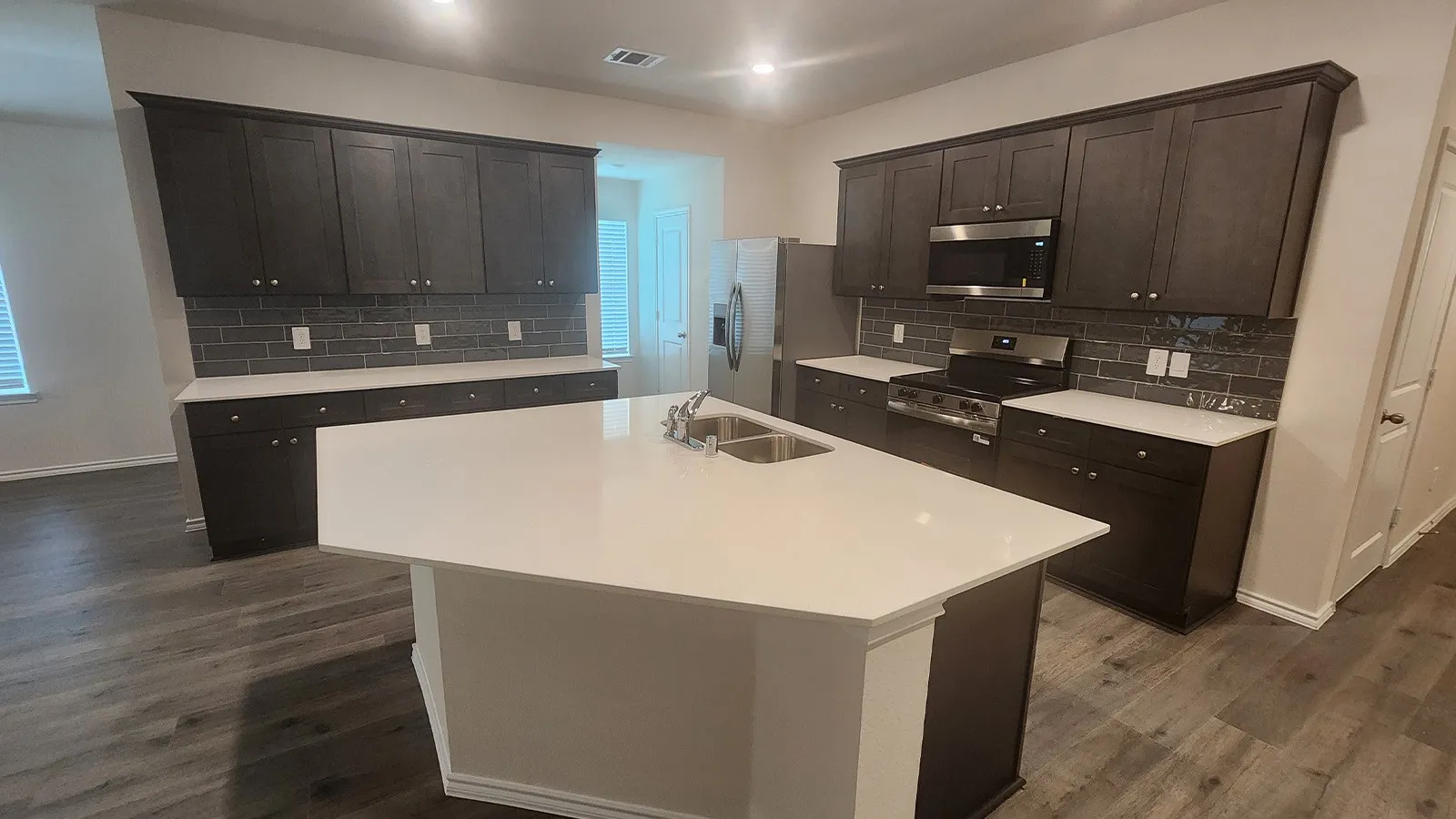 Modern kitchen featuring an island with a double basin sink, stainless steel appliances, dark wood-finish cabinetry, white countertops, and a gray subway tile backsplash