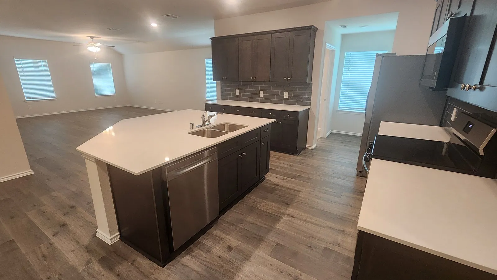 Kitchen featuring an expansive island with a stainless steel double basin sink, dark wood-finish cabinetry, light-toned countertops, a subway tile backsplash, and wood-finish flooring