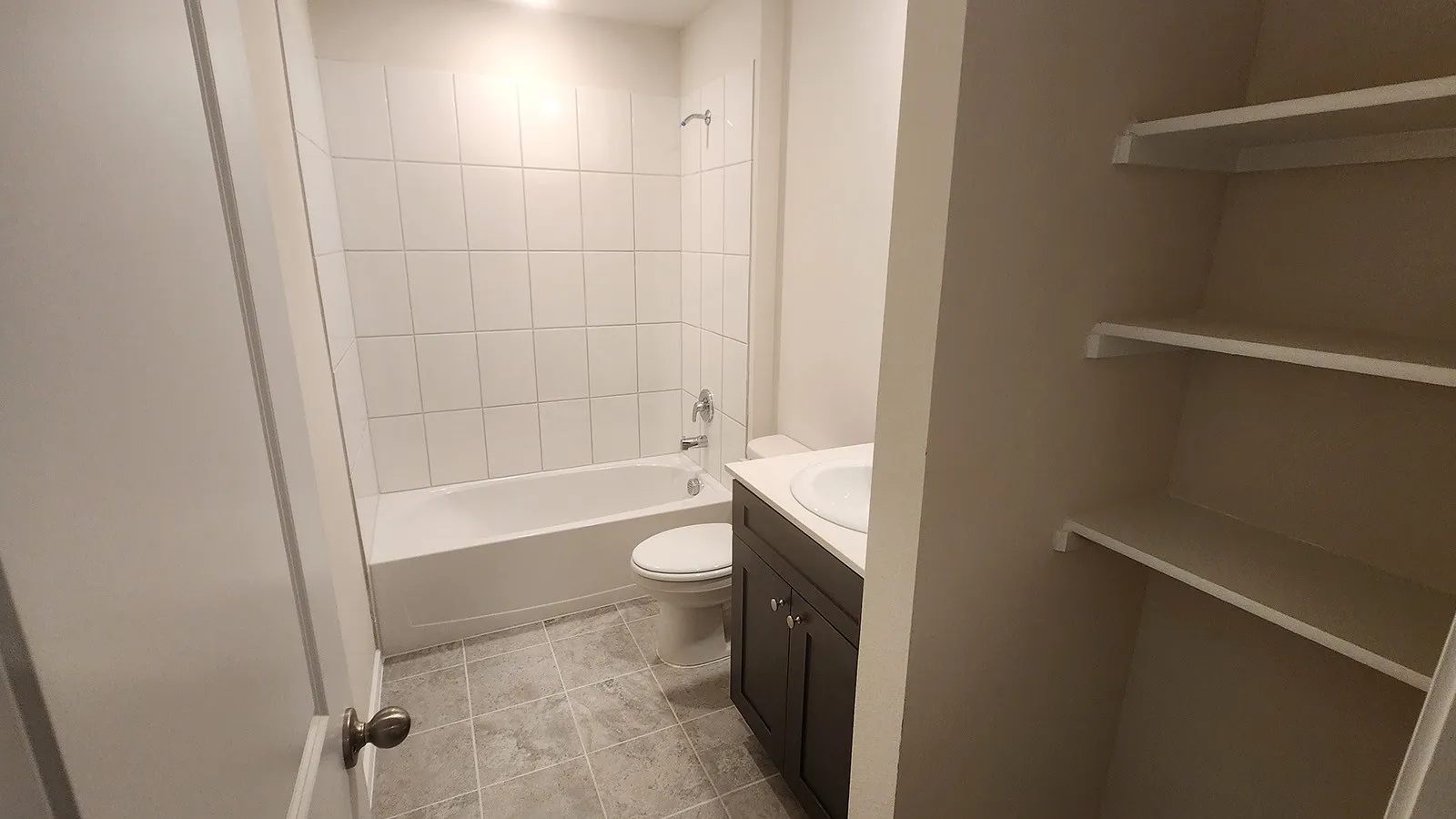 Bathroom featuring a white tub, white tiled shower surround, dark wood-finish vanity, white countertop with integrated sink, and built-in shelving