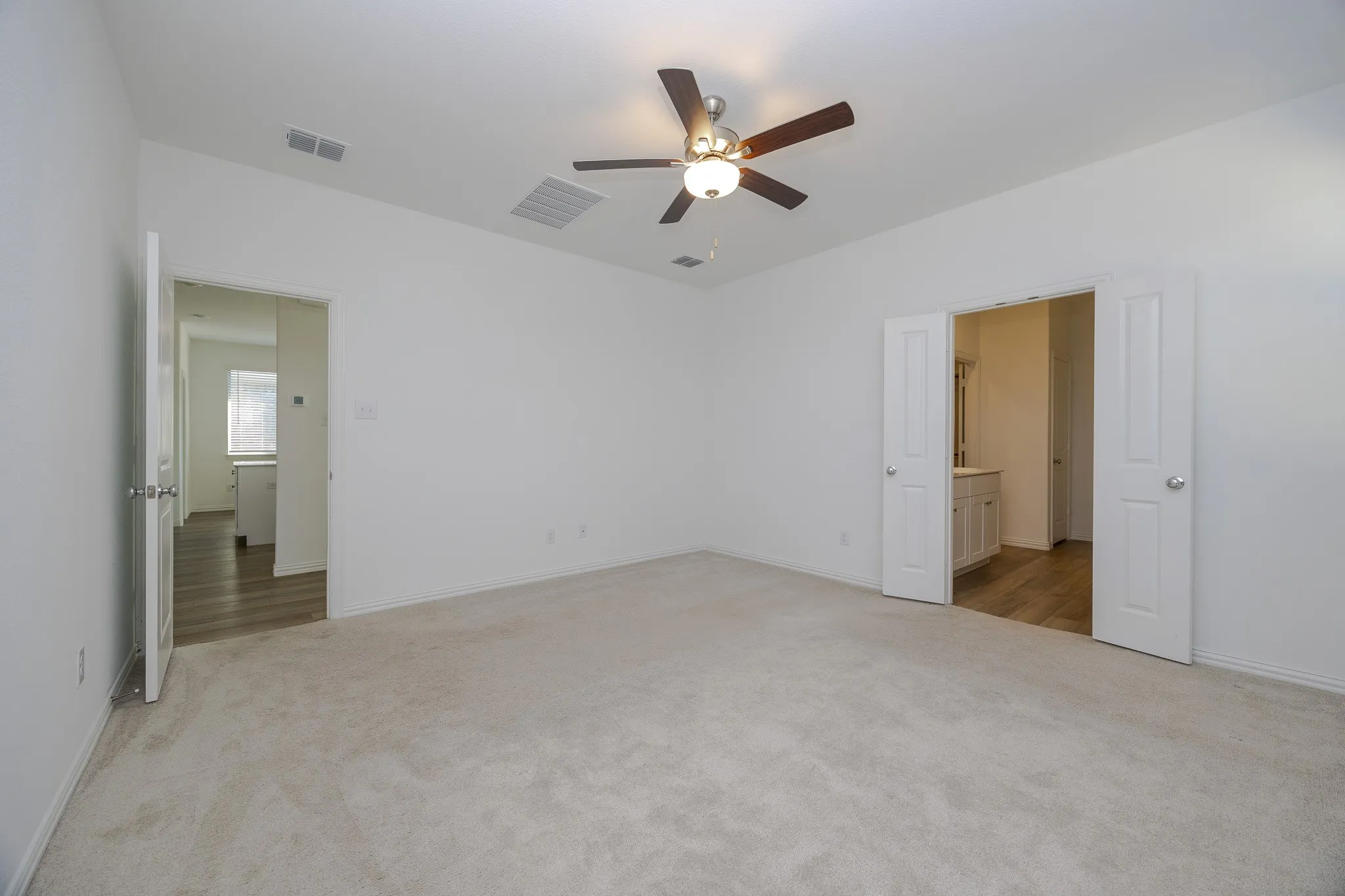 Carpeted room with a ceiling fan light fixture and dual white paneled doors leading to a space with wood-finish flooring and a vanity