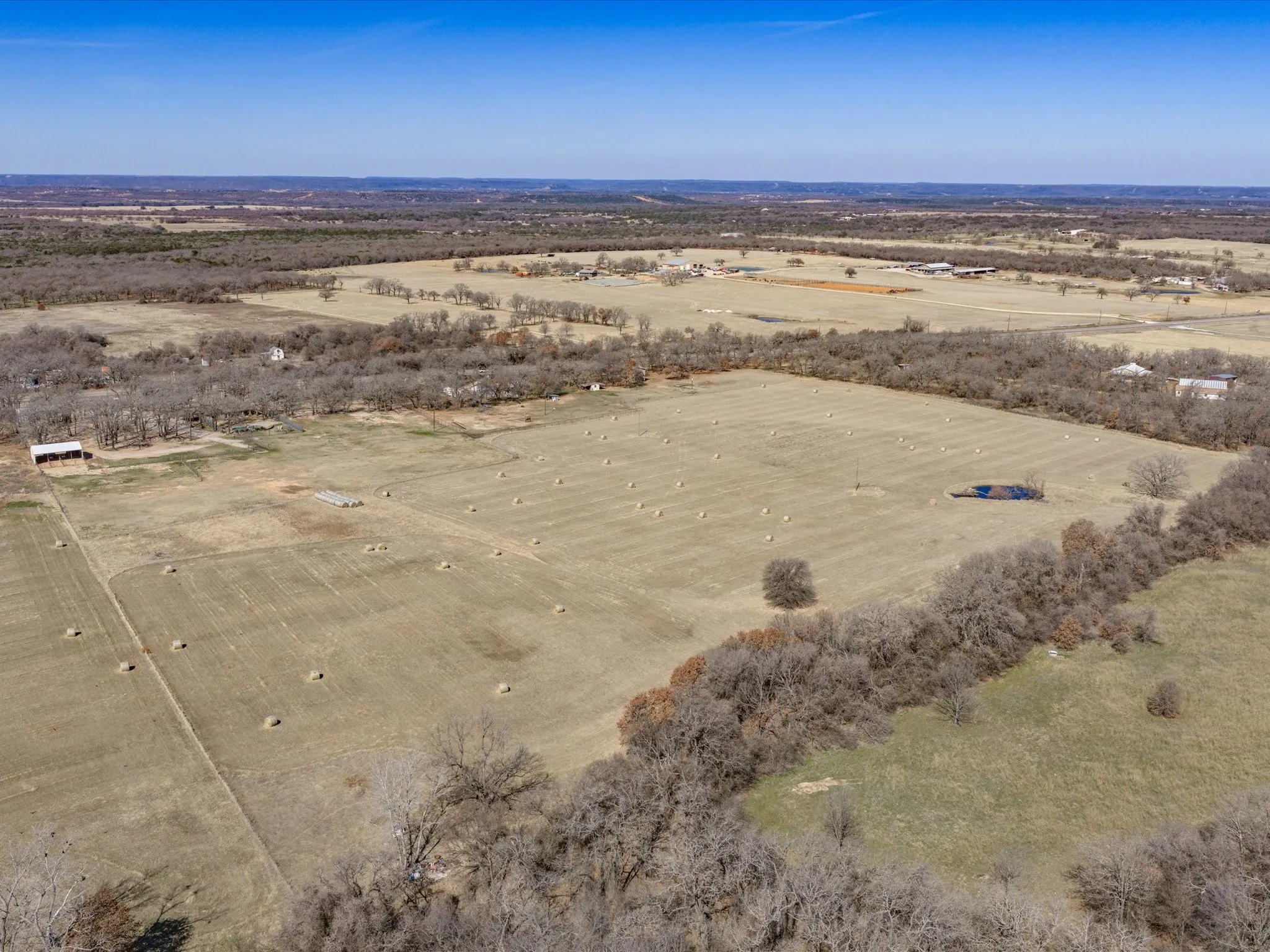 Expansive agricultural land featuring a natural pond, scattered hay bales, and perimeter tree lines